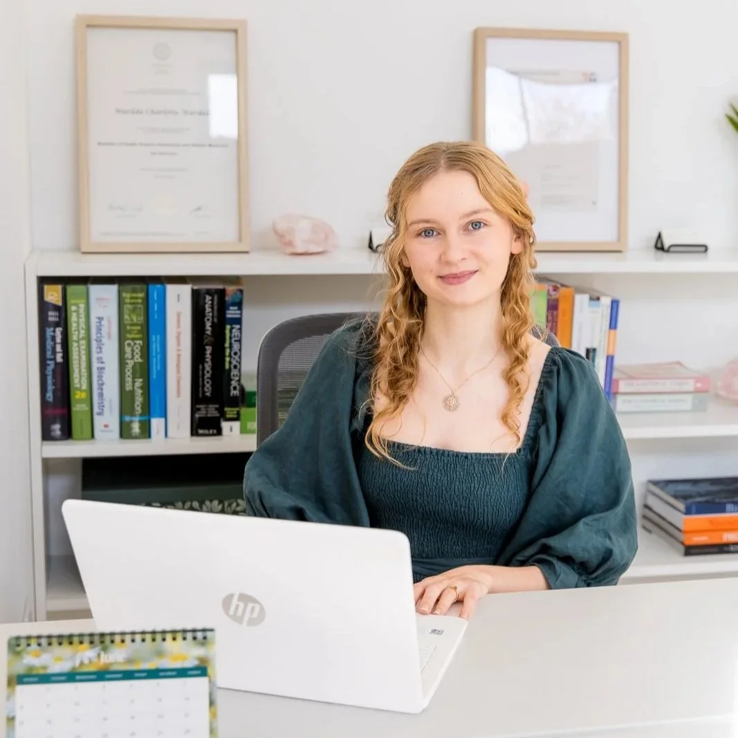 A woman with long curly blonde hair sitting at a desk with a white HP laptop, in an office with bookshelves and framed degrees on the wall behind her.