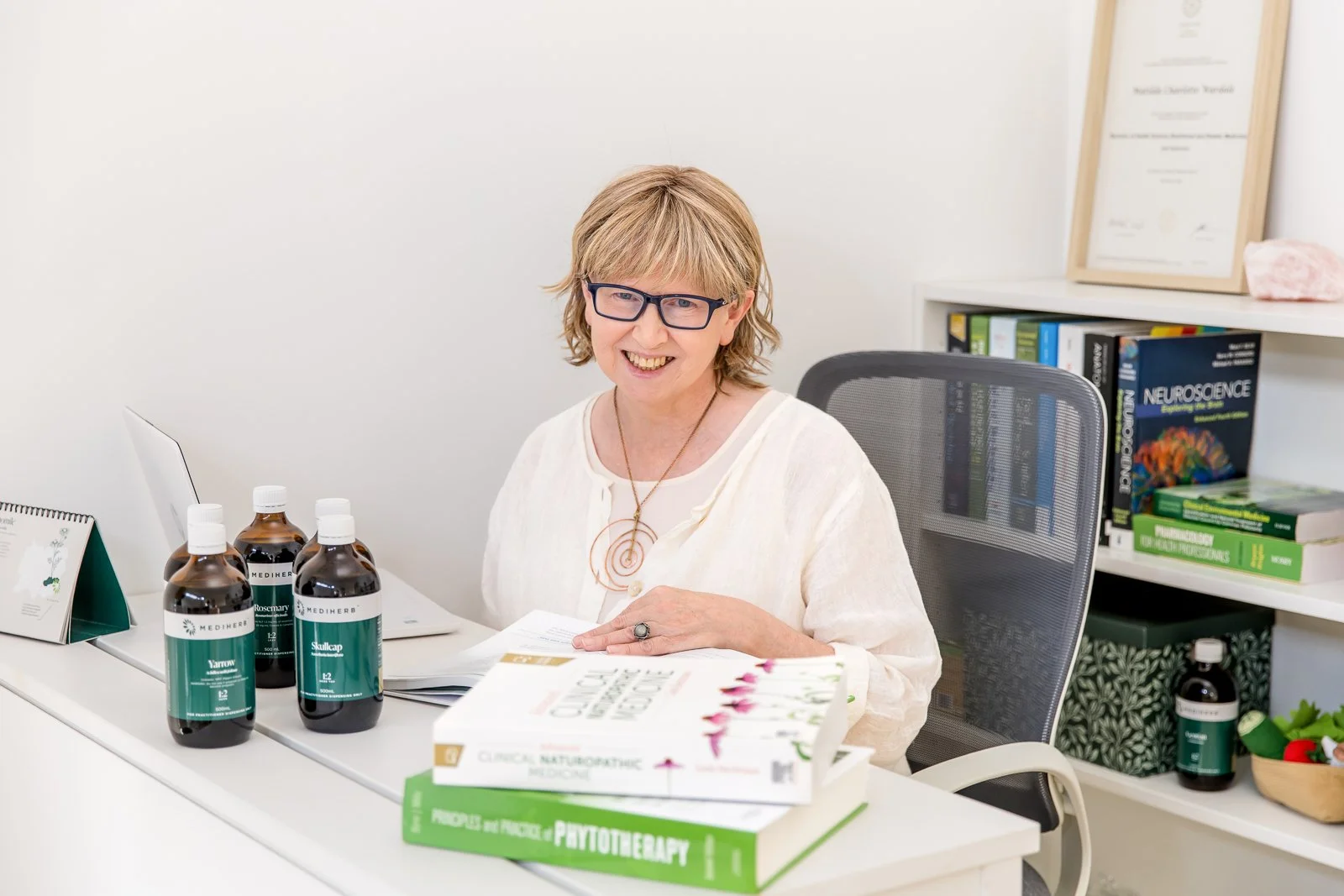 A woman with glasses sitting at an office desk, smiling, with books, bottles, and papers on the desk, and a bookshelf with medical textbooks and a framed certificate behind her.