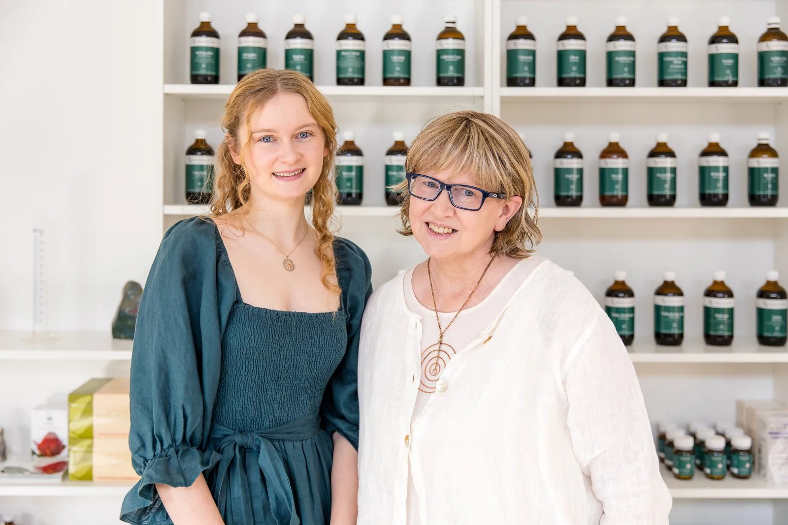 Two women smiling in a herbal medicine clinic with shelves of brown jars with herbs in the background.