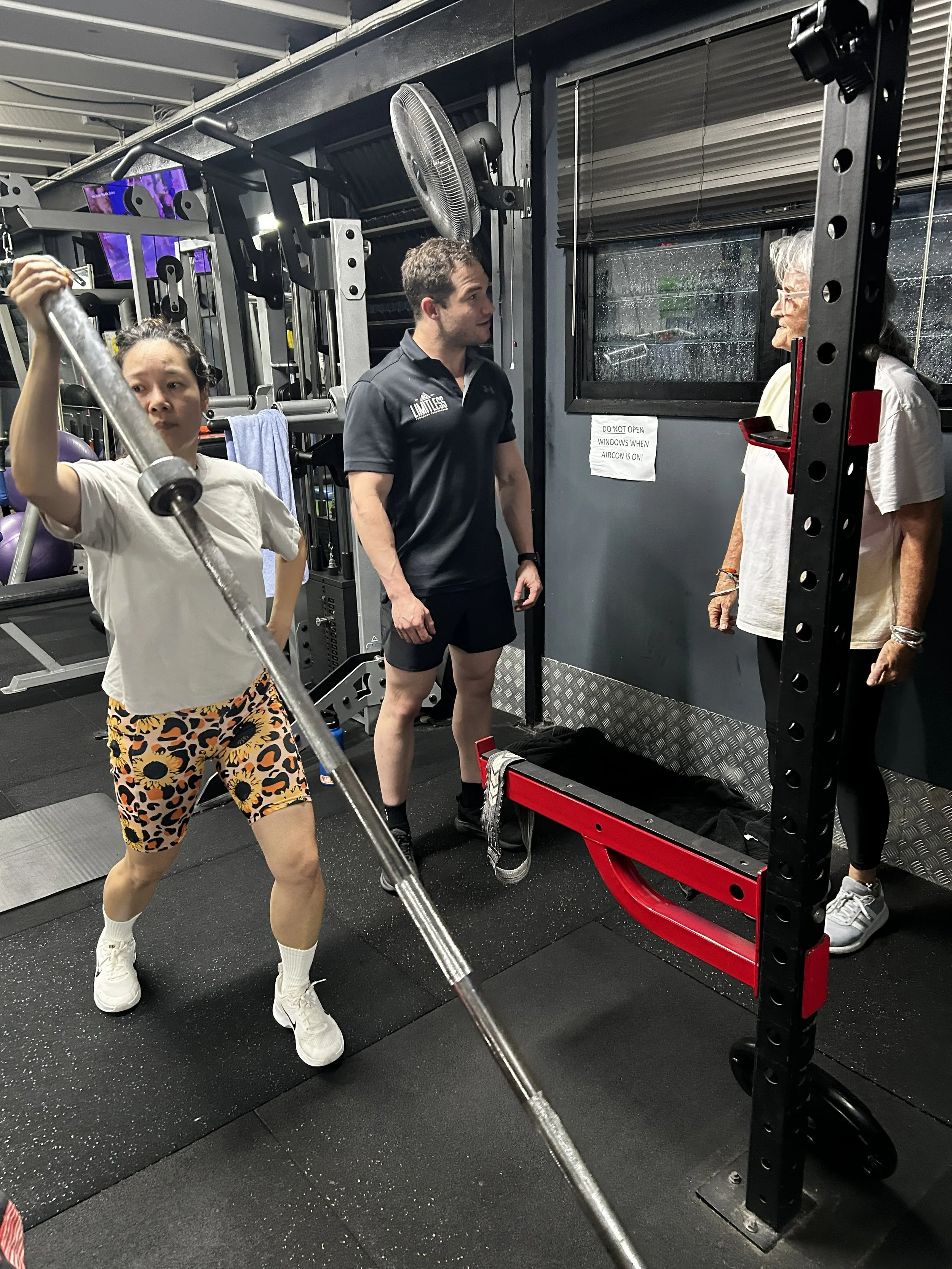A woman is lifting a barbell at the gym, wearing leopard print shorts and a white shirt, with a man and an elderly woman engaging in conversation nearby. The gym has black flooring, wall-mounted TVs, and gym equipment in the background.