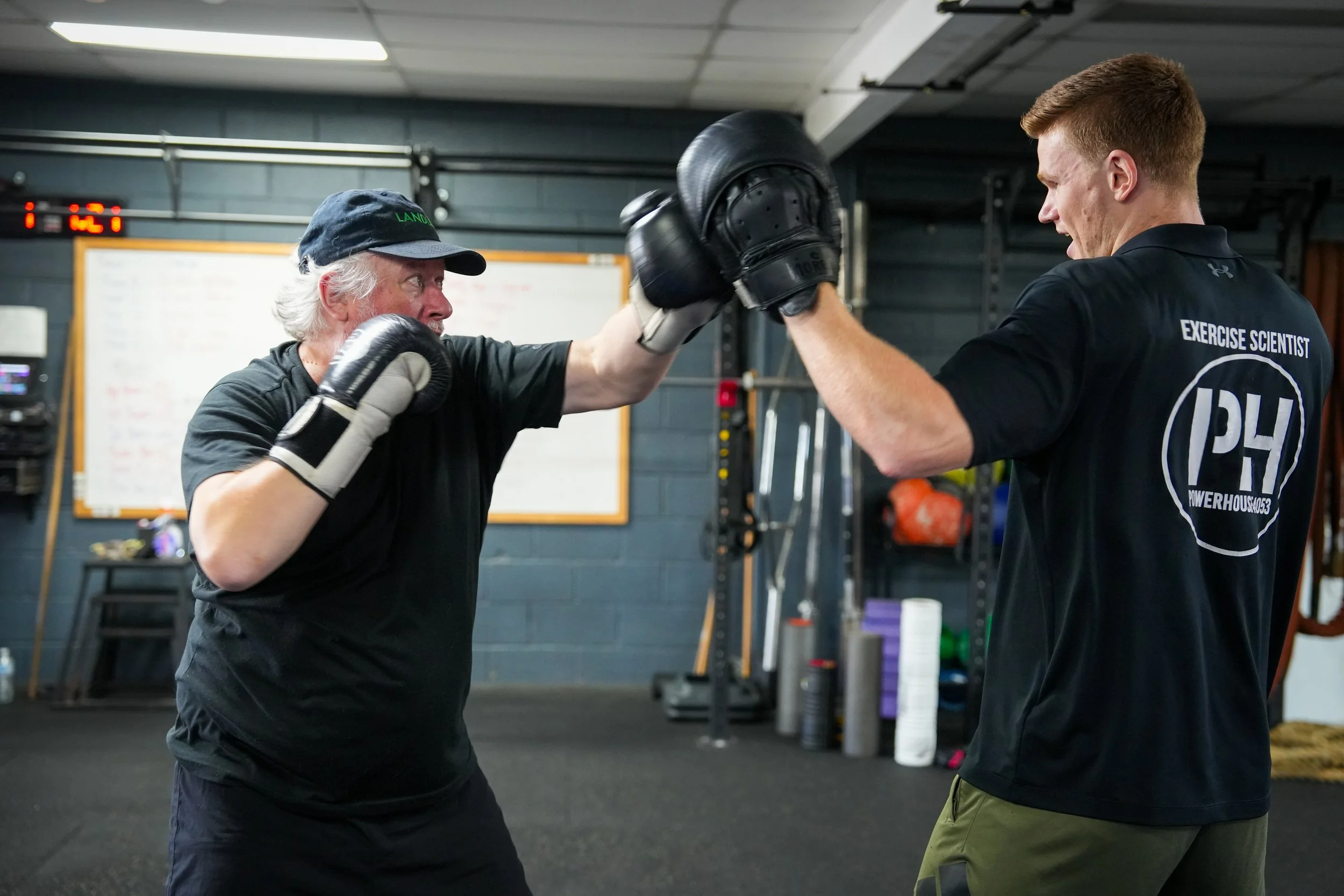 An older man and a younger man are practicing boxing in a gym, with both wearing gloves, and touching gloves in a training session.