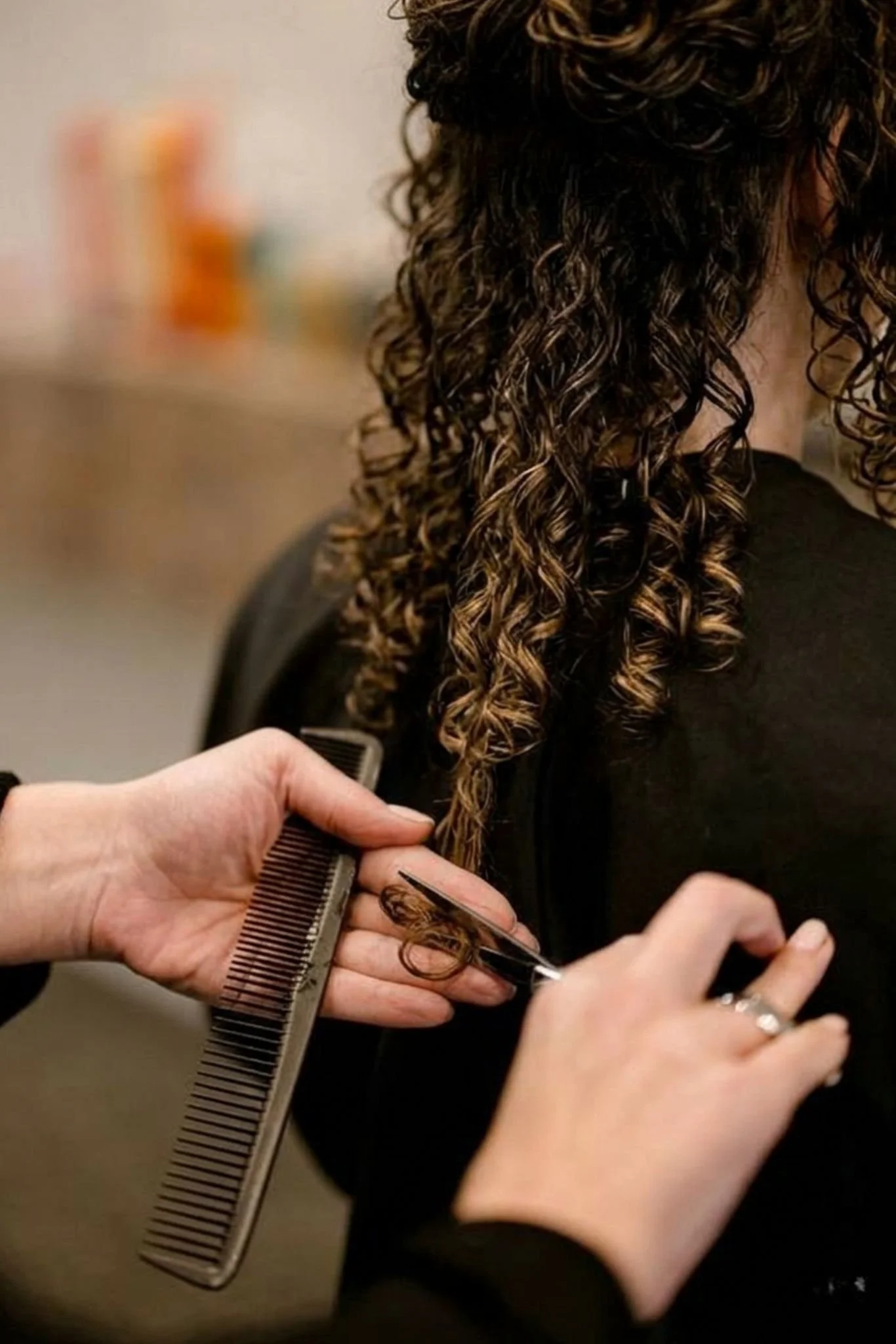 A person having their curly hair styled, with a hairstylist trimming the ends using scissors and holding a wide-tooth comb, in a salon setting. Curly Haircut.