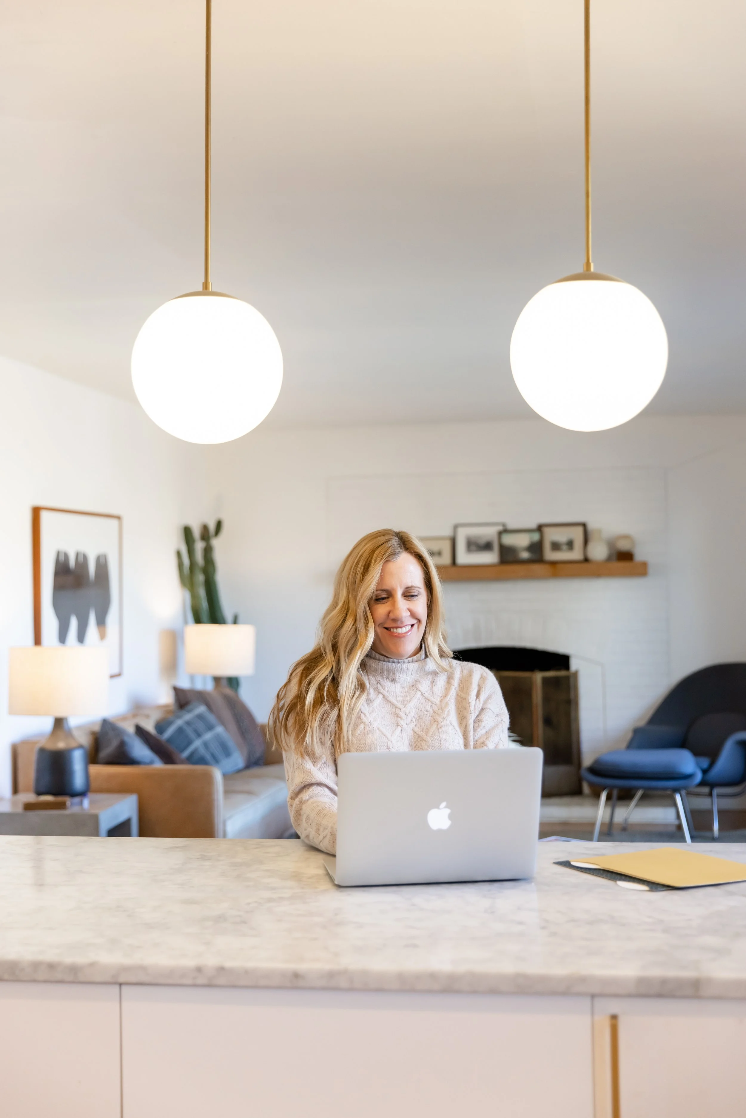 A woman smiling while working on a silver Apple MacBook laptop at a kitchen island in a modern home interior.
