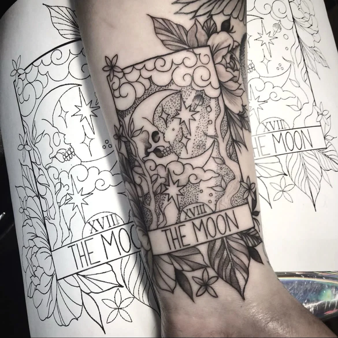 A tattoo design on a person's forearm featuring the tarot card 'The Moon,' with the number XVIII and a banner at the bottom reading 'The Moon.' The design includes clouds, stars, a crescent moon, and a skull surrounded by flowers and leaves, with lin