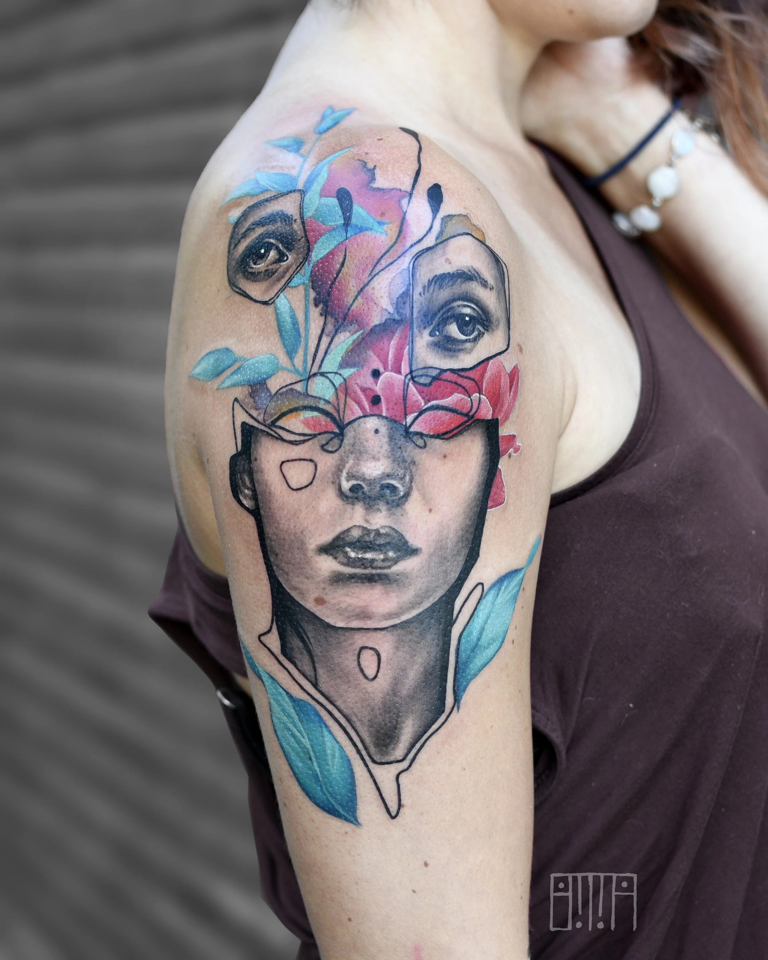 A tattoo on a woman's upper arm featuring a woman's face with watercolor-style flowers, leaves, and abstract elements blending into her head, with two realistic eyes above her face and colorful accents.