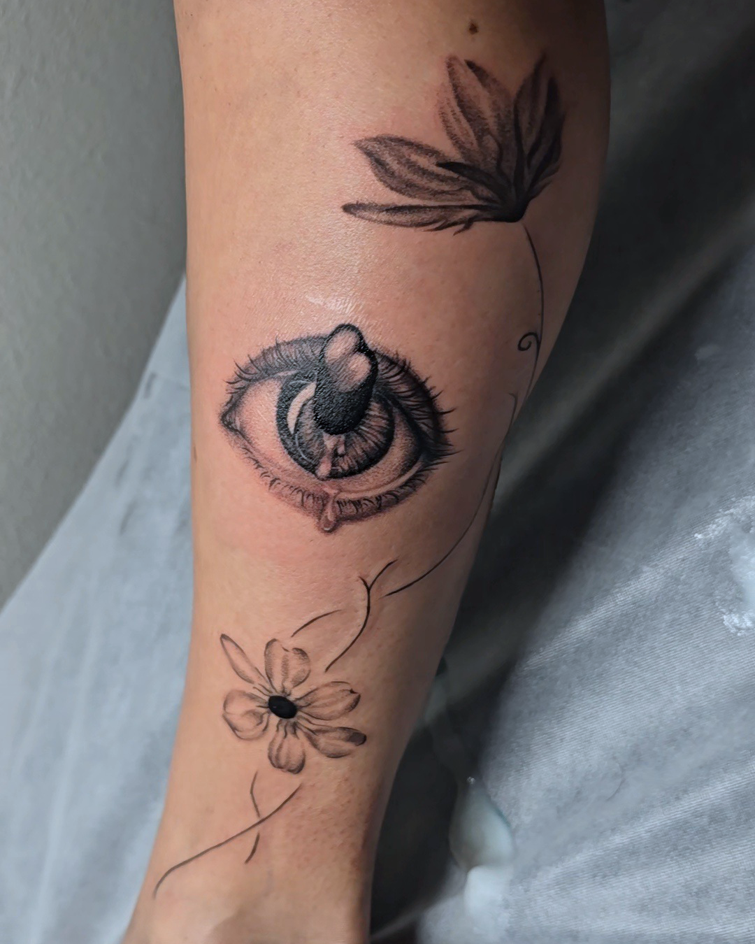 A leg tattoo of an eye with long eyelashes, a tear drop, and a thin stemmed flower.
