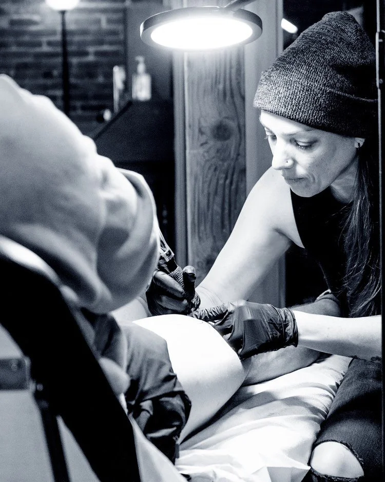 Tattoo artist working on a client's leg under bright lighting, wearing gloves and a beanie hat.