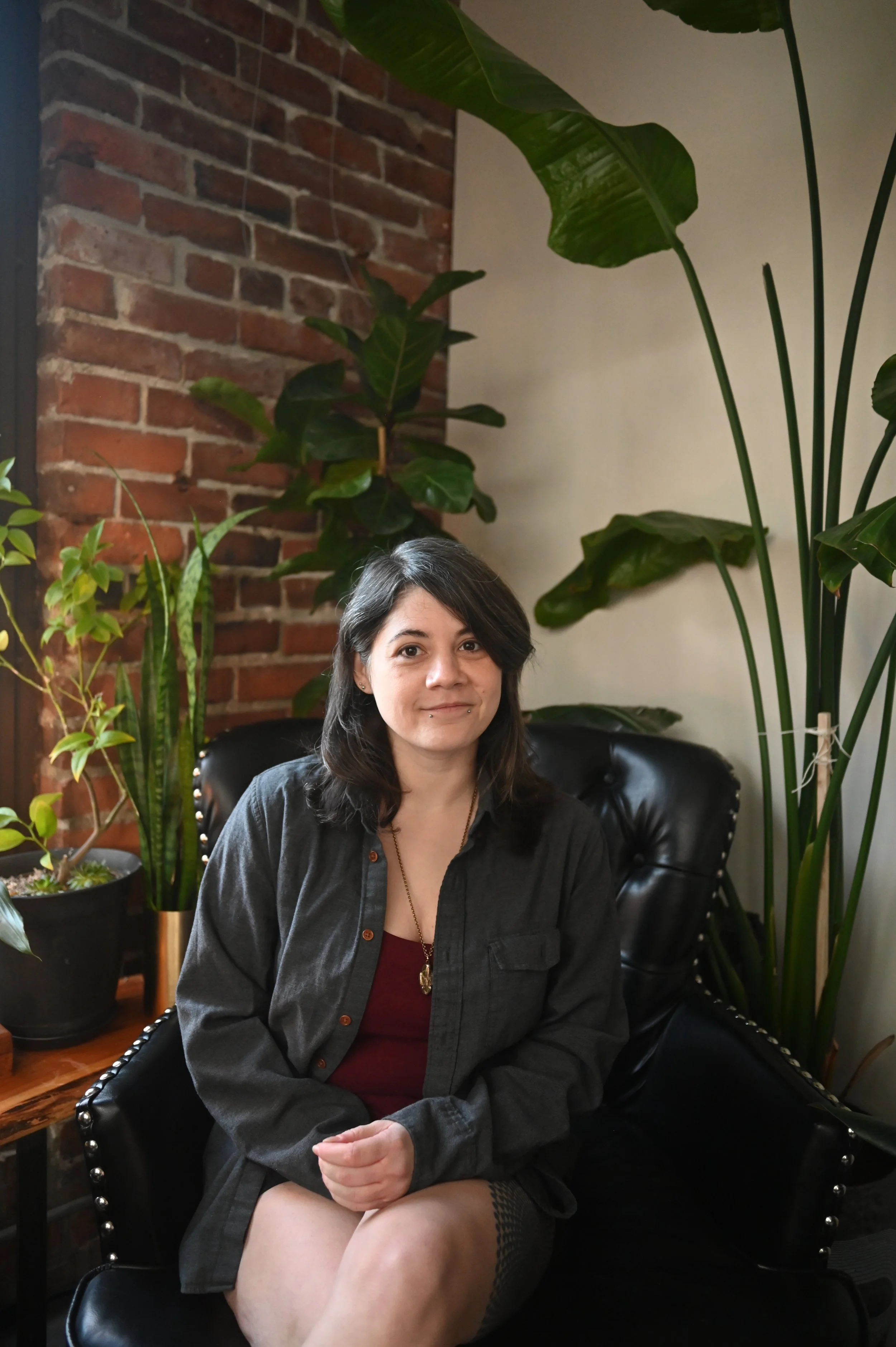A woman with dark hair sitting on a black leather armchair surrounded by tall green houseplants in a room with a brick wall and a plain light-colored wall.