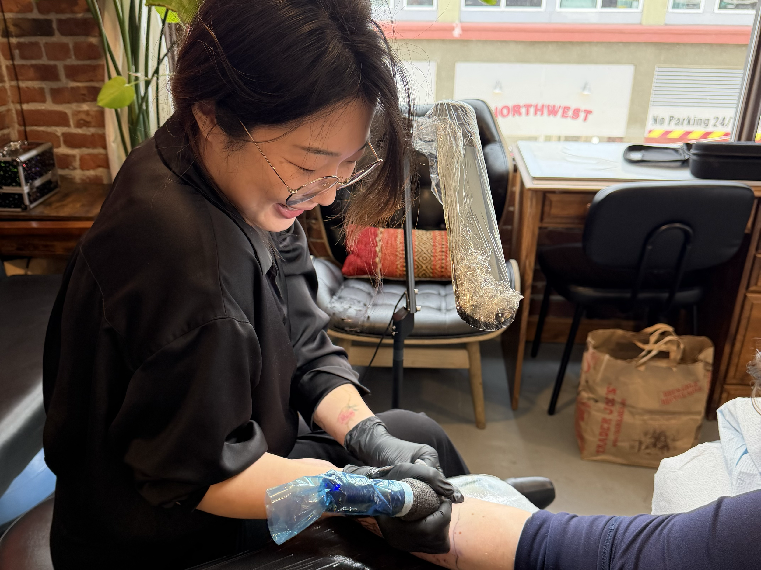 A woman with long dark hair, glasses, and tattoos on her arm, wearing black gloves, tattooing someone's arm in a tattoo studio with brick walls, potted plants, and a window showing a building outside.