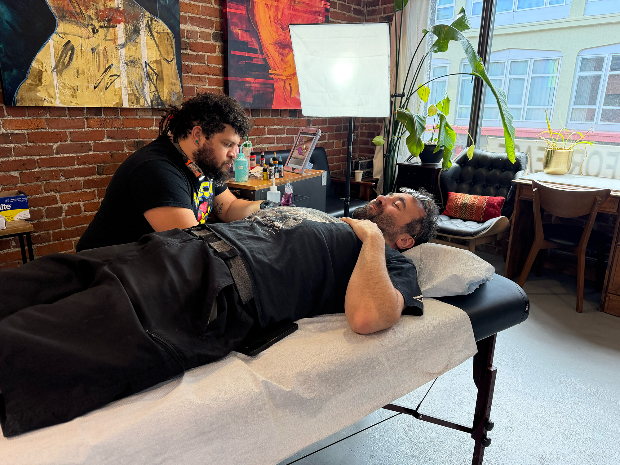 A tattoo artist working on a tattoo on a man's arm in a bright, art-filled studio. The studio has exposed brick walls adorned with colorful abstract paintings, large windows with natural light, and potted plants near the window.