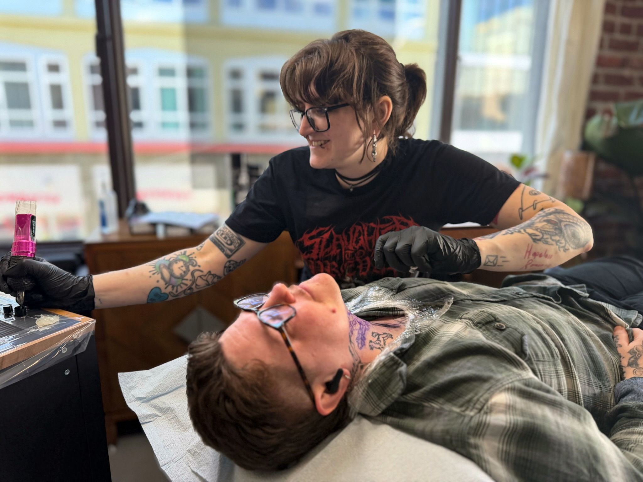 A person with tattoos and glasses smiling as they work on a tattoo on a person's arm in a tattoo studio.