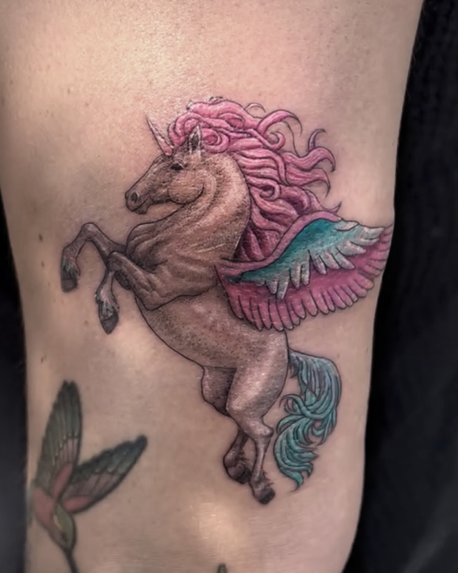 A realistic tattoo of a unicorn with pink hair and blue and pink wings on a person's skin.