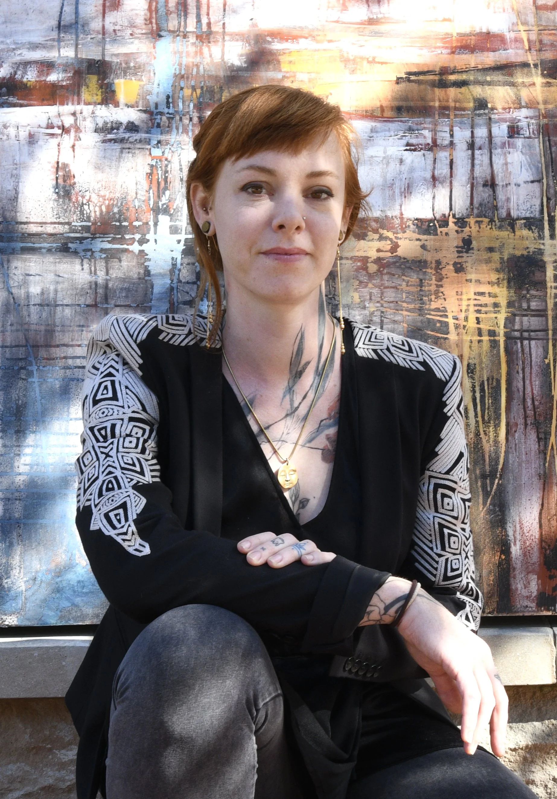 A woman with short red hair, tattoos on her chest and hands, and multiple earrings, sitting in front of abstract graffiti art on a wall. She is looking at the camera with a slight smile.