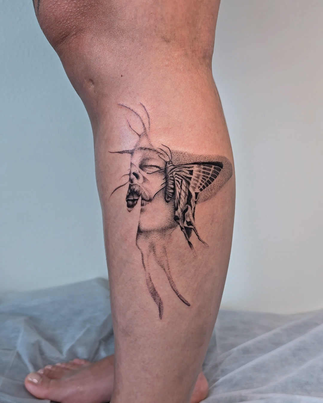 Tattoo of a dragonfly with a human face, featuring detailed wings and antennae, on a person's calf.
