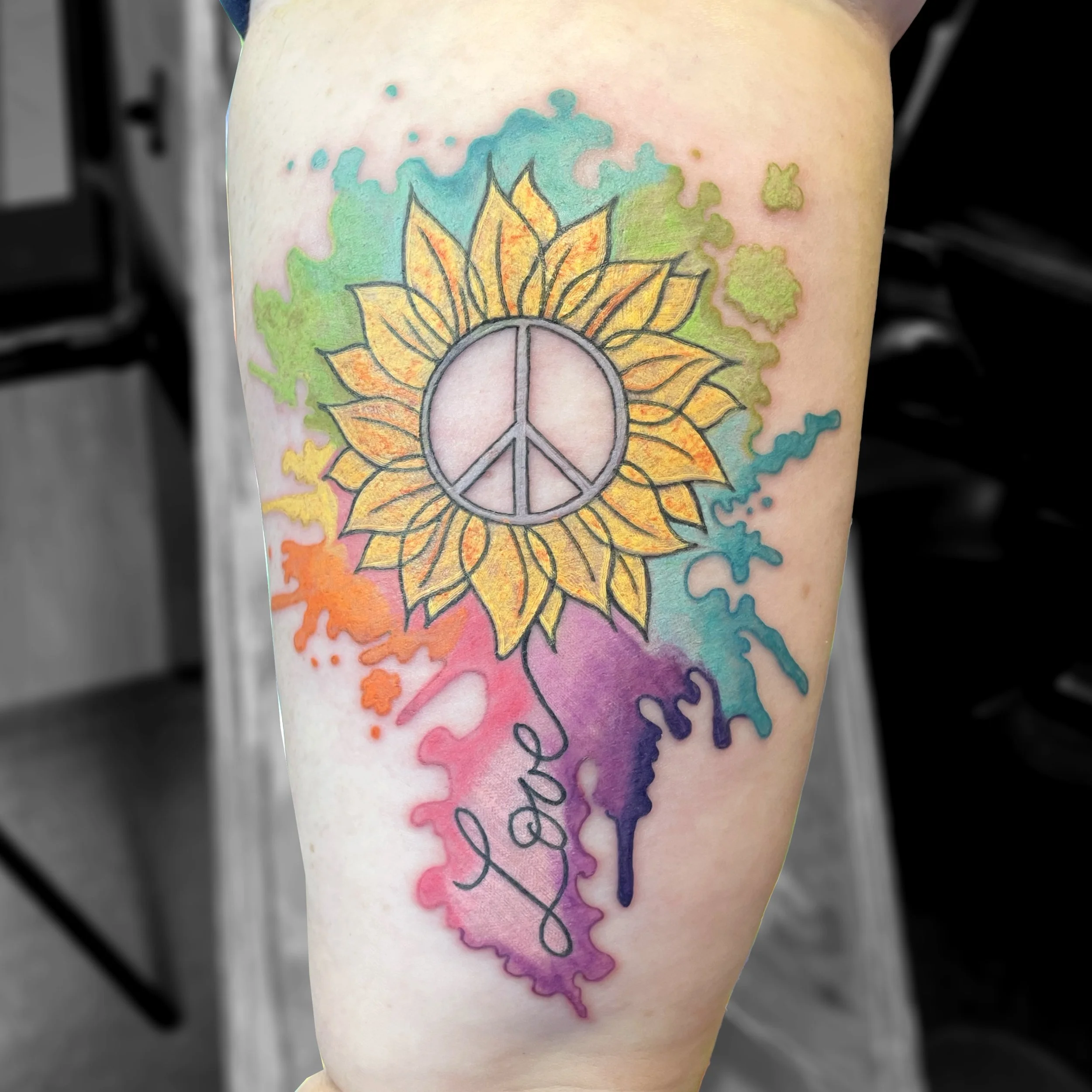 Colorful tattoo of a sunflower with a peace symbol in the center, surrounded by watercolor splashes in rainbow colors and the word 'love' written in cursive.