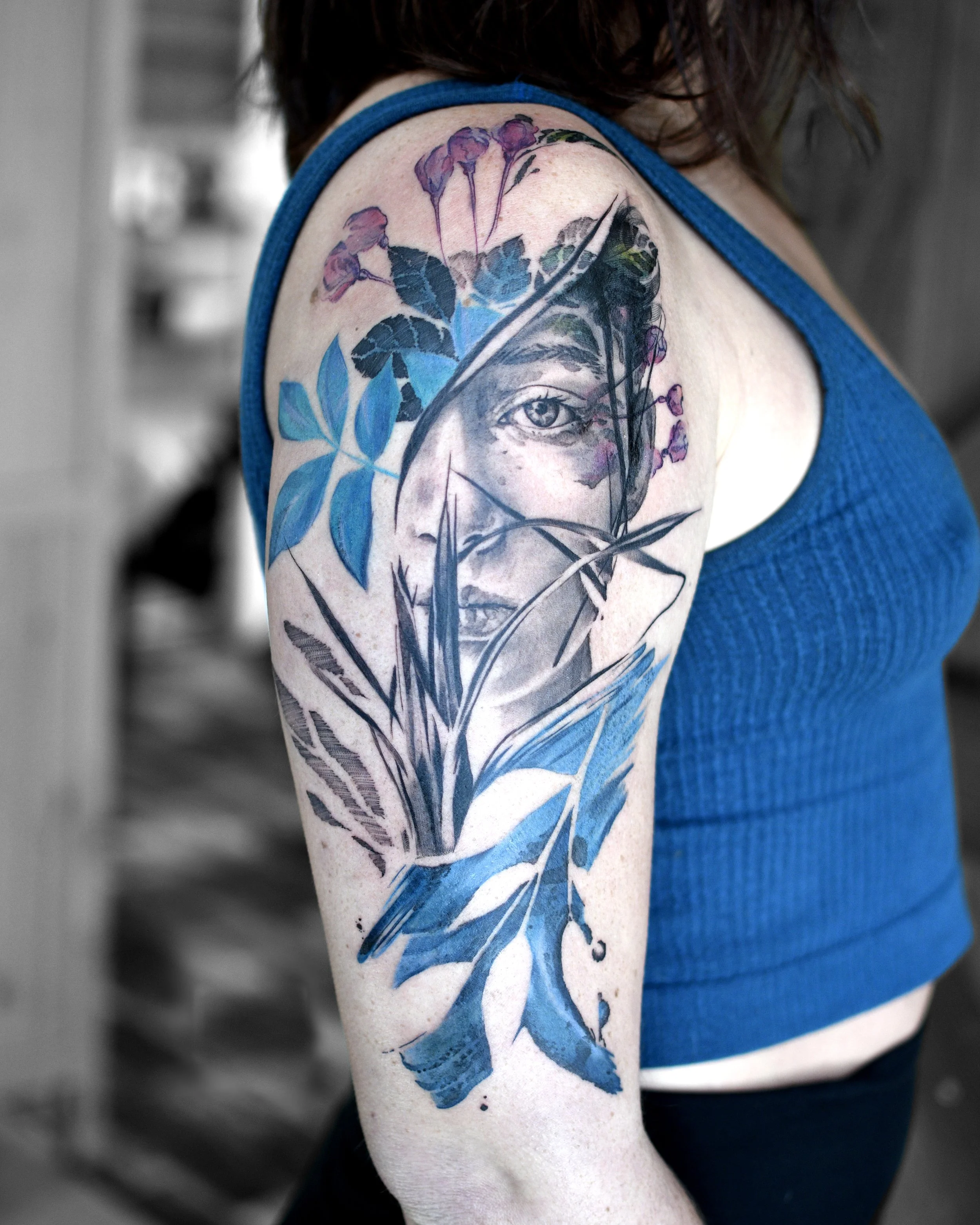 Tattoo of a woman's face partially obscured by leaves and branches in a painterly style on upper arm .
