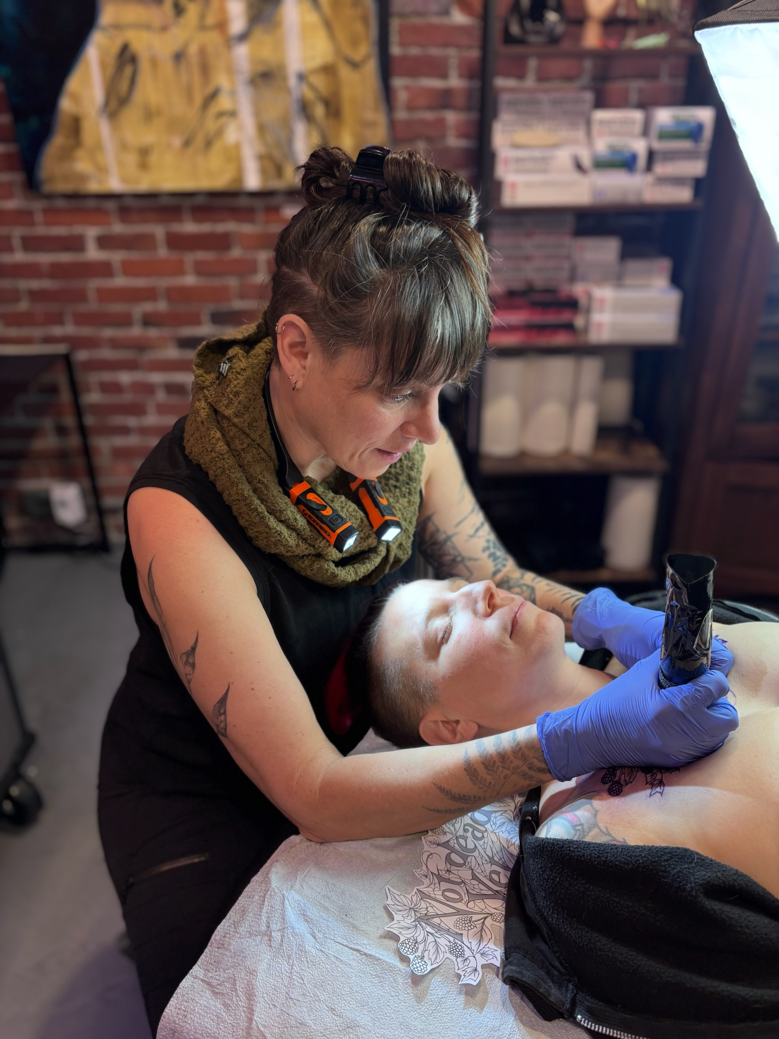 Tattooed woman with short hair, wearing a green scarf, giving a tattoo to a person lying on a table in a tattoo studio with brick walls.