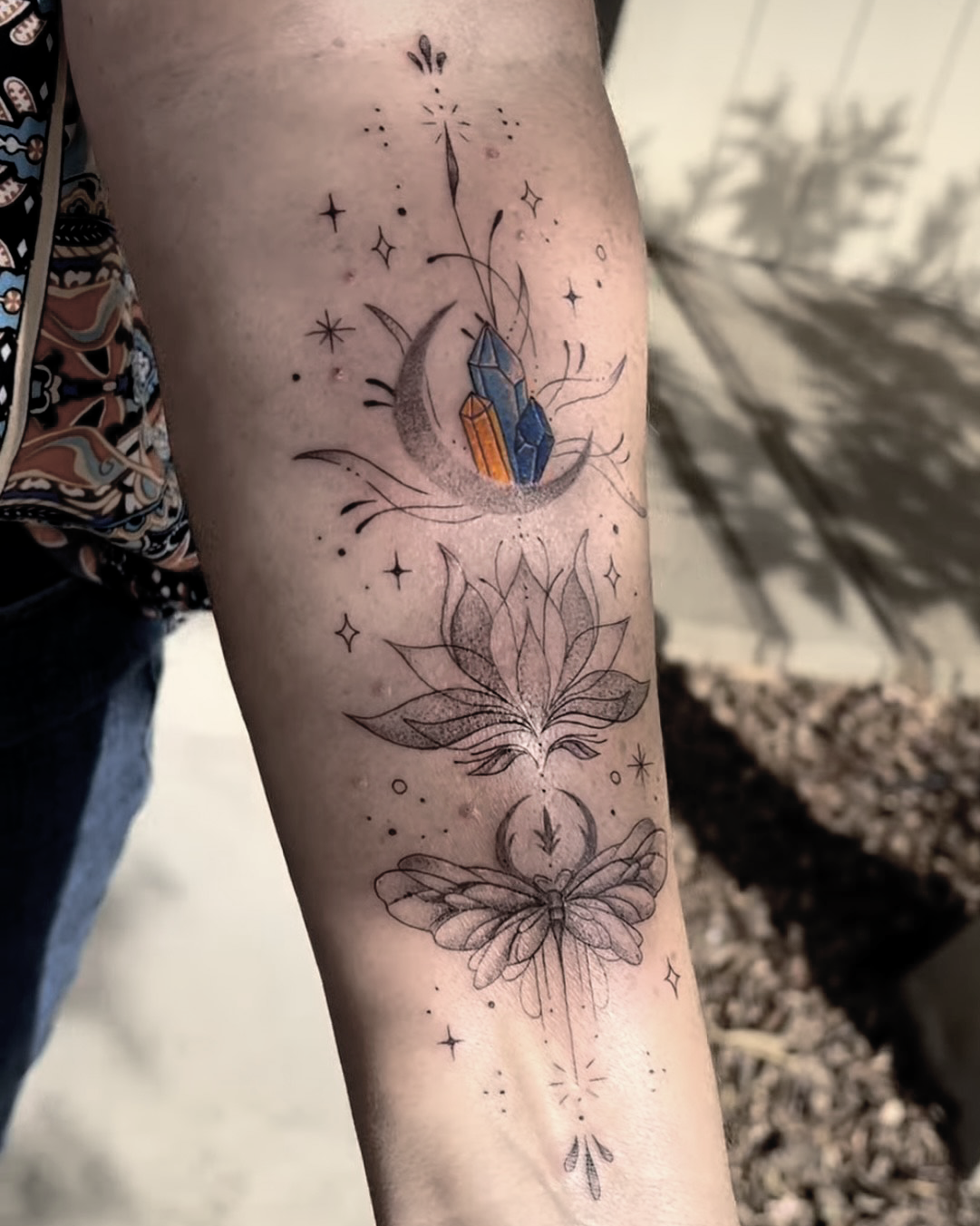 A fine line tattoo of a geometric crystal cluster on a crescent moon, a lotus flower, and a butterfly with stars and celestial elements, in black ink with some colored accents.