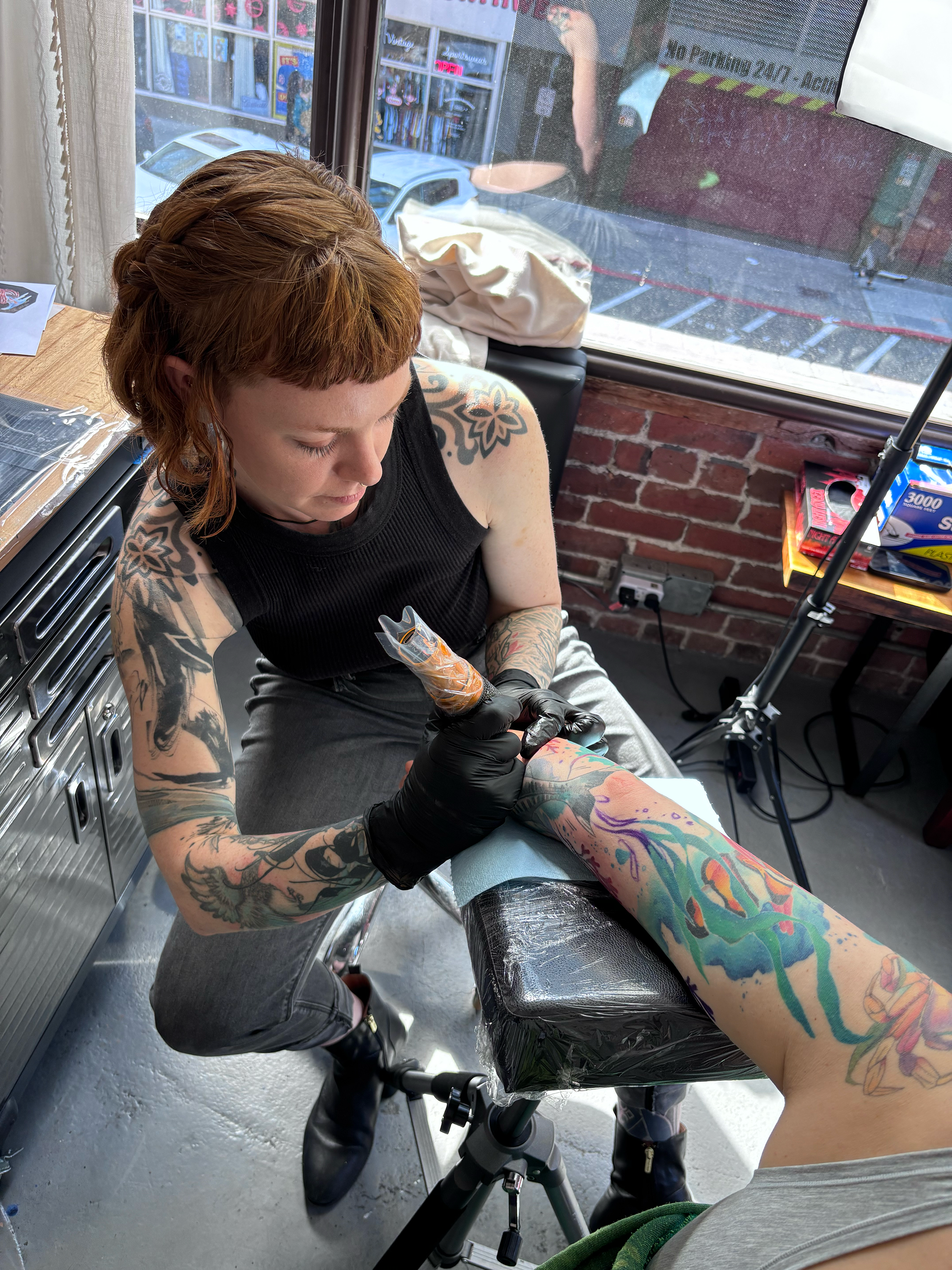 A tattooed woman getting a tattoo on her arm at a tattoo shop, with a person standing nearby in the reflection of the window.