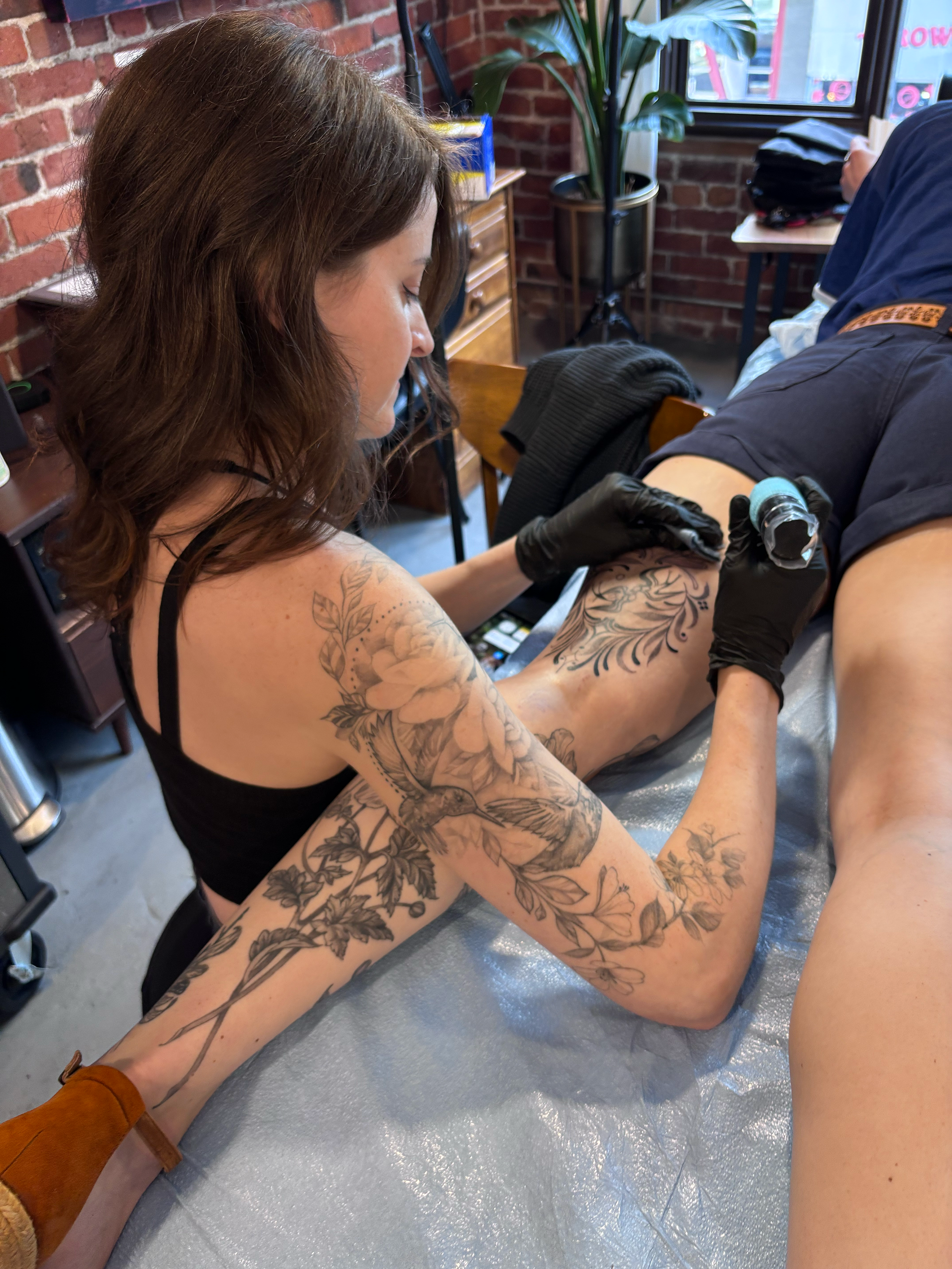 A woman with tattoos on her arms and legs is sitting in a tattoo shop, receiving a tattoo on her upper thigh from a tattoo artist. The shop has a brick wall and a large window.