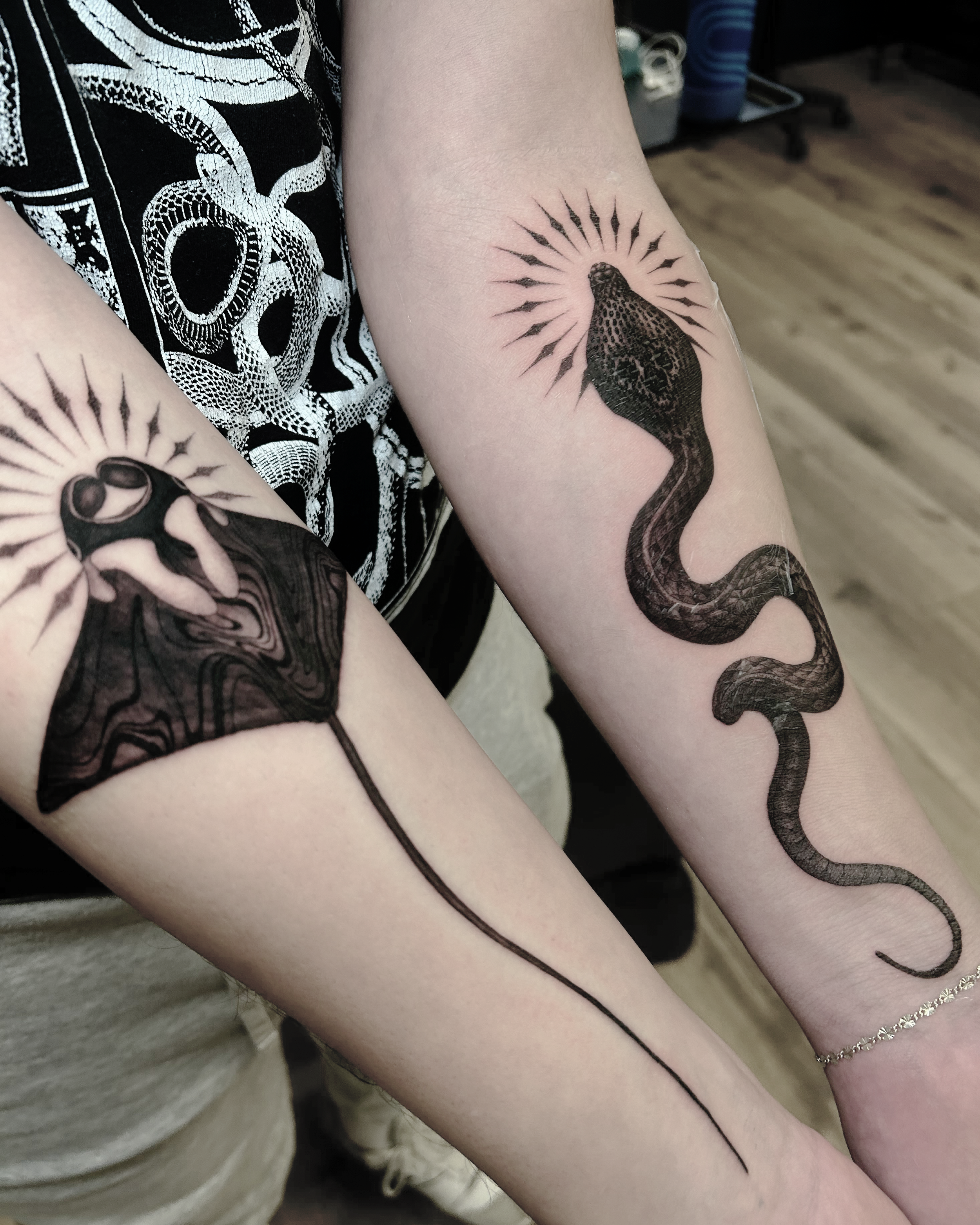 Two arms with high detail blackwork tattoos: one tattoo of a cobra with a sunburst halo, and the other of a manta ray with a sunburst halo.