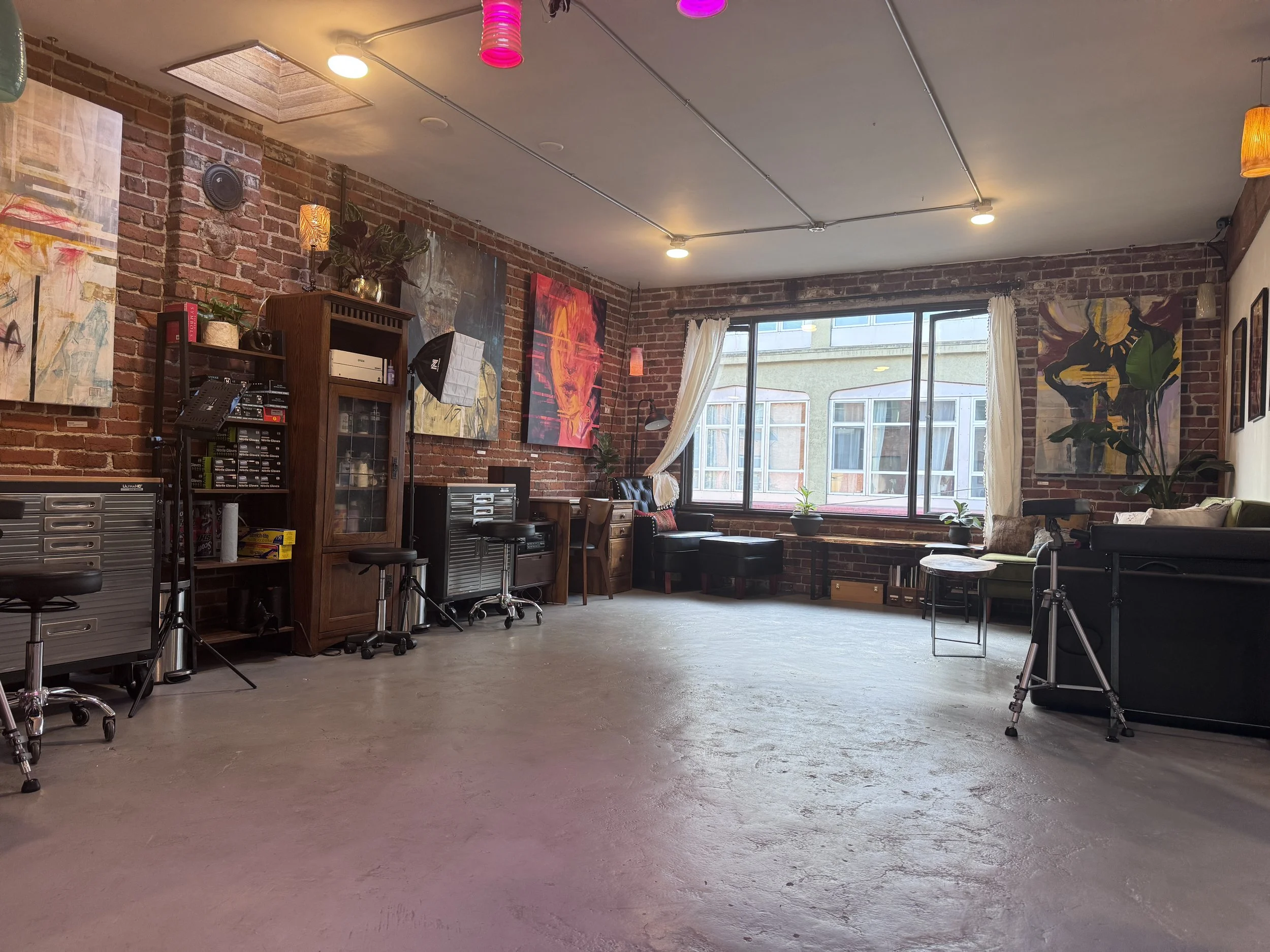 A spacious art studio with exposed brick walls and large windows letting in natural light. The room contains several colorful abstract paintings on the walls, a wooden cabinet, chairs, and some plants. Studio equipment like stands and lighting are also visible.