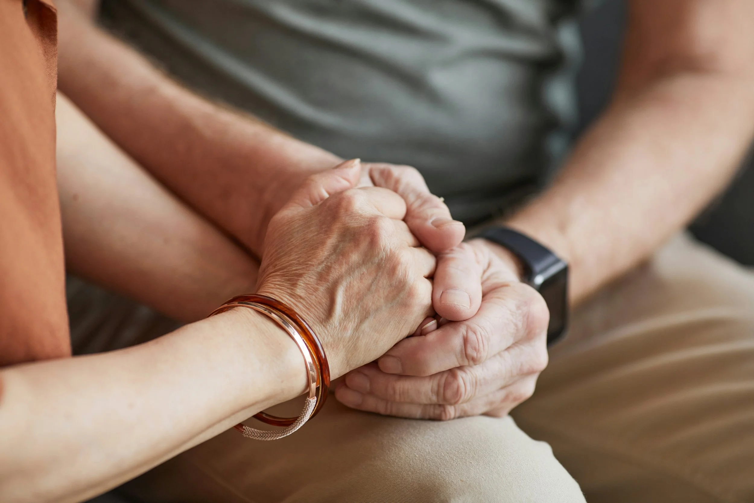 Close-up of two hands clasped together, one with a bracelet, resting on a person's arm. The background shows parts of two people, one wearing a gray shirt and the other wearing a brown bracelet.