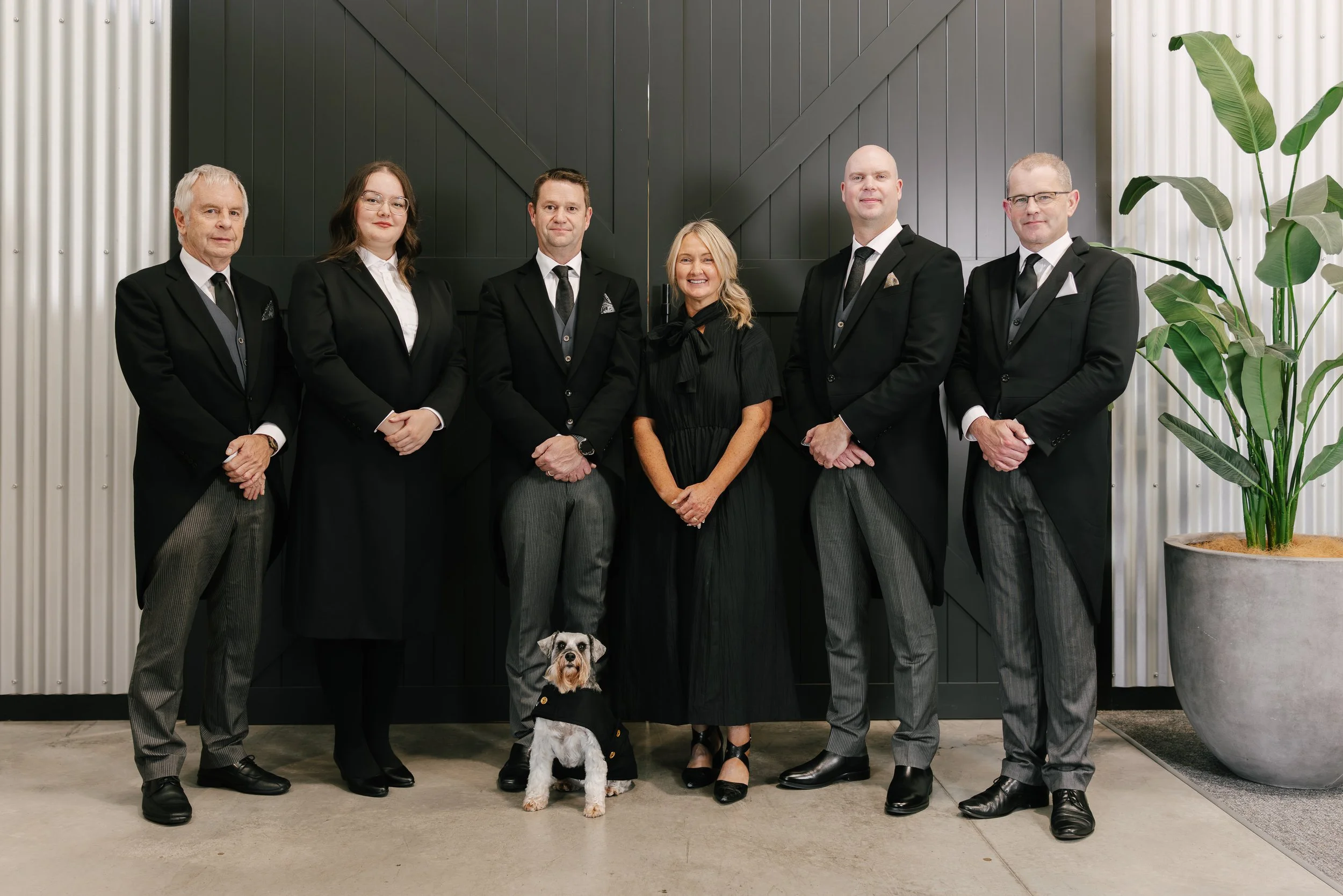 Group of six people in formal black suits, white shirts, and black shoes standing together in front of a modern black wall with potted plants on either side, and a small dog in a black coat sitting in front of them.