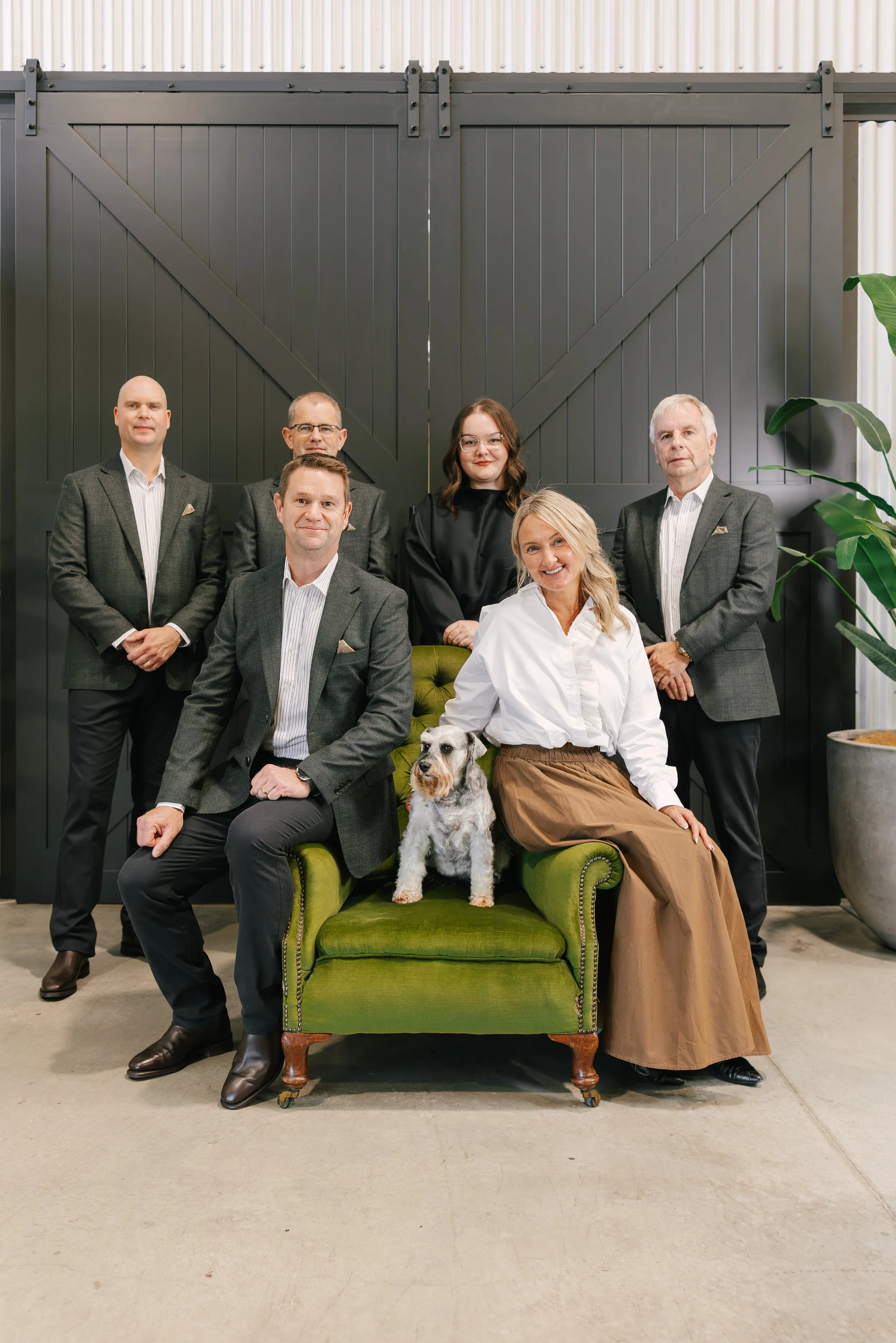Group portrait of six people in business attire, with a woman and dog seated on a vintage green loveseat, in front of a large black barn door and industrial background.