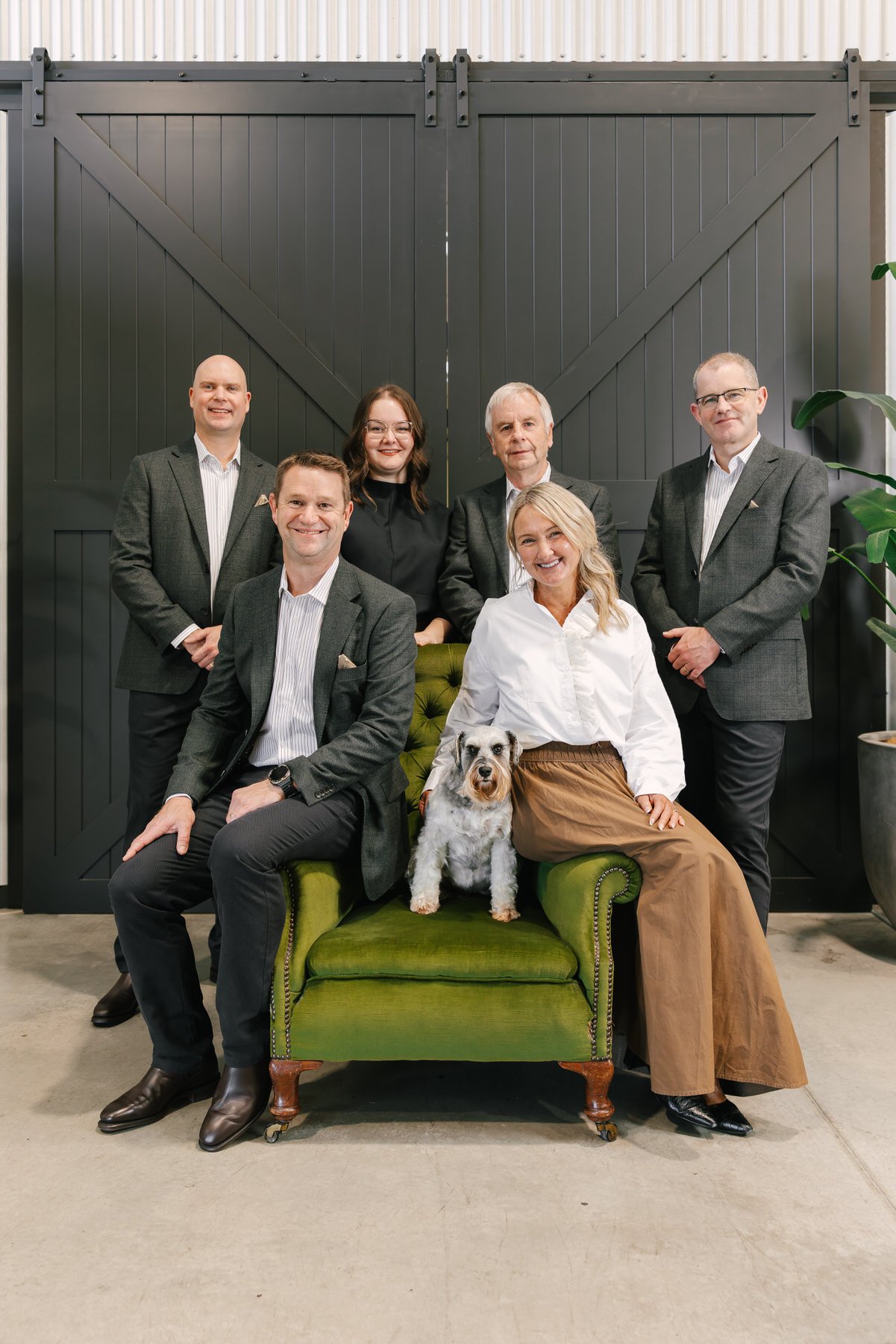 Group of six adults in business attire posing with a dog on a green vintage sofa in front of a black barn door.
