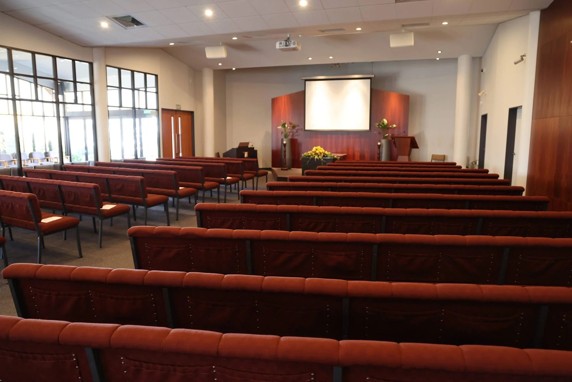 Empty conference room with red upholstered chairs arranged in rows, a large projection screen at the front, and decorative vases with flowers on pedestals on either side of the screen.