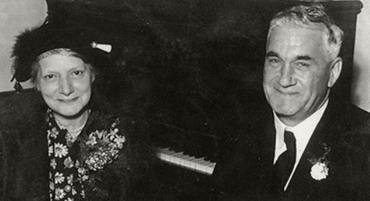 Black and white photo of a woman and a man sitting together, the woman wearing a hat and floral dress, and the man wearing a suit and tie, both smiling.