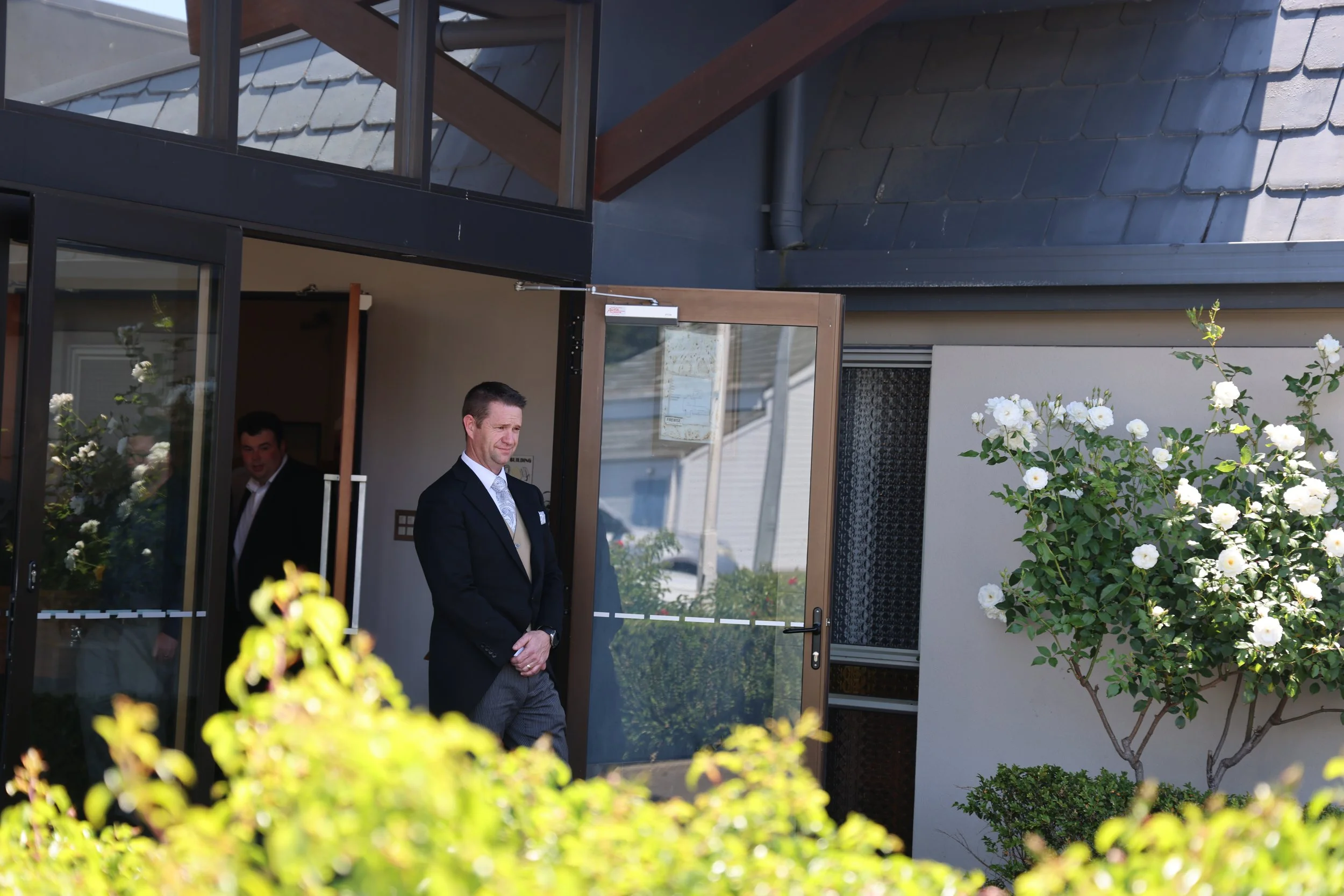 Two men in formal suits are standing near the entrance of a modern building with glass doors. One man is in the foreground, smiling slightly, while the other is partially visible inside.