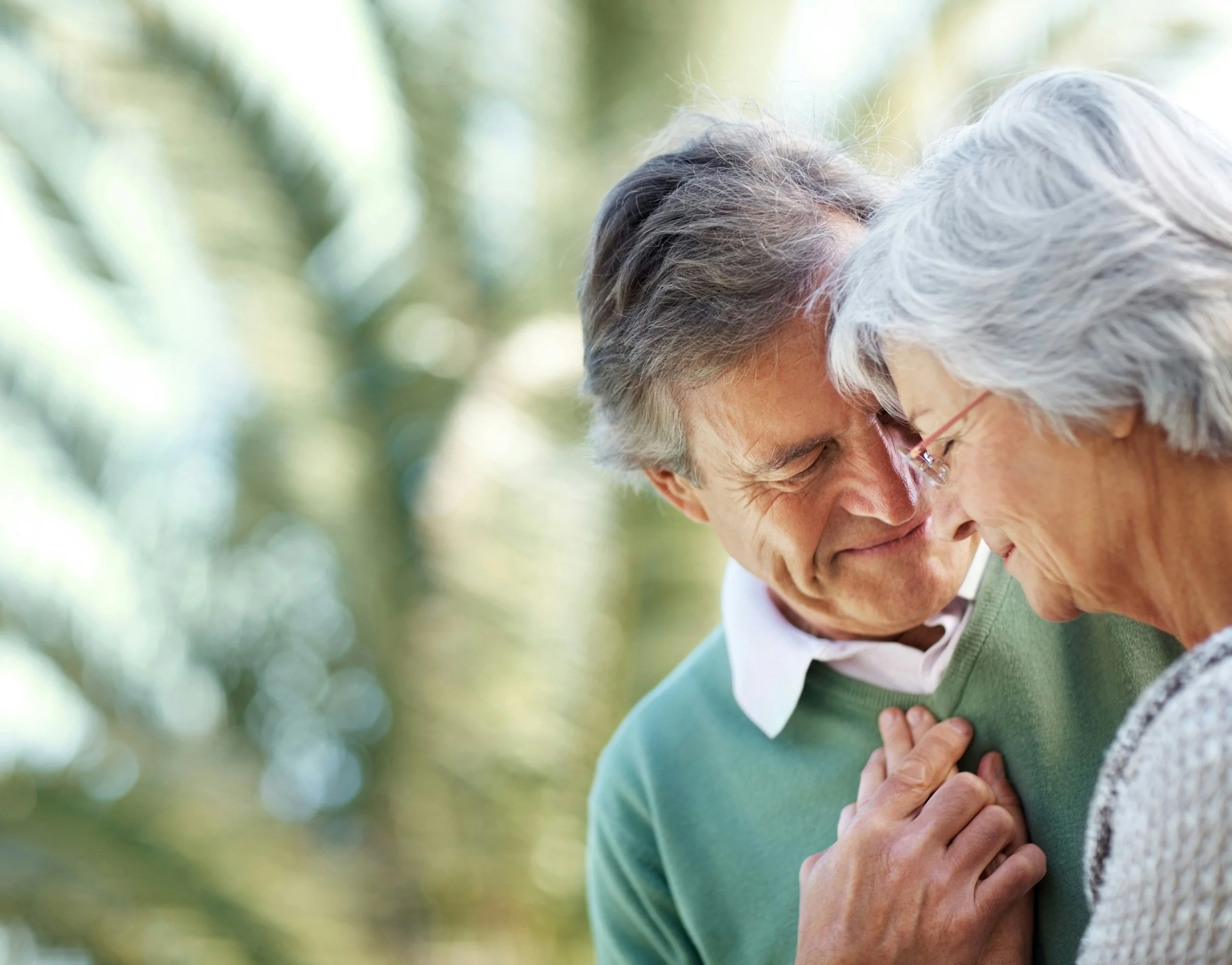 An elderly couple with gray hair touching foreheads and expressing affection outdoors near greenery.