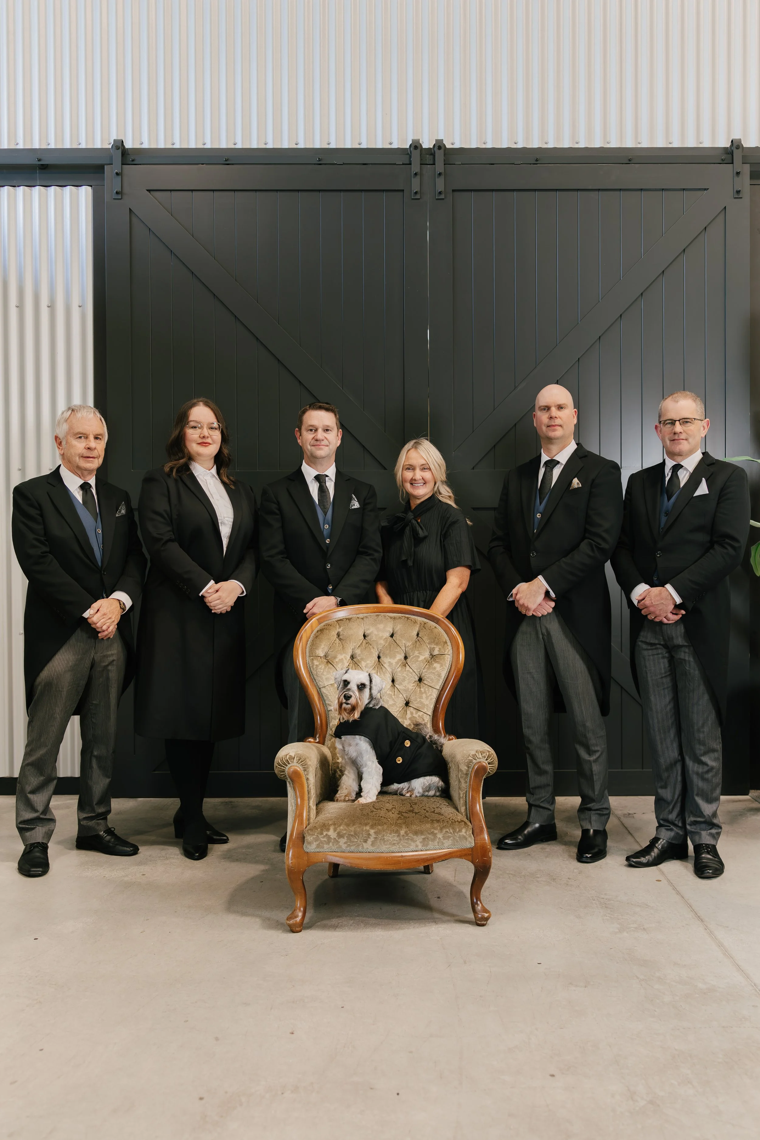 Six people in formal attire standing in a row behind a vintage armchair with a dog sitting on it, in front of a large black barn door and corrugated metal wall.