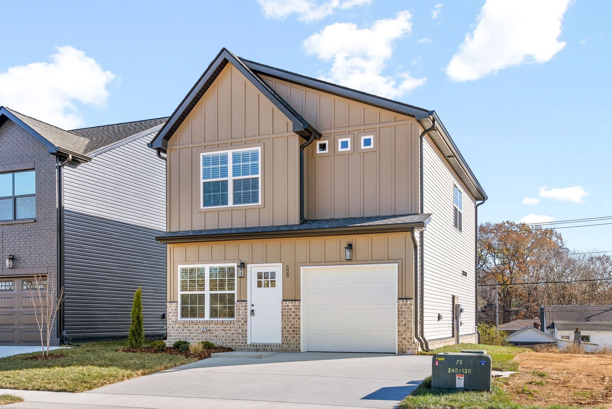 New two-story house with tan siding, brick accents, a white garage door, and a white front door, situated in a suburban neighborhood with a small lawn and driveway.