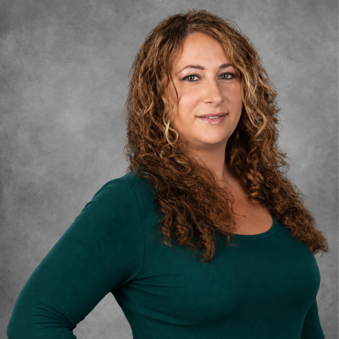 Portrait of a woman with curly red hair and green eyes, wearing a purple top, standing against a dark gray background.