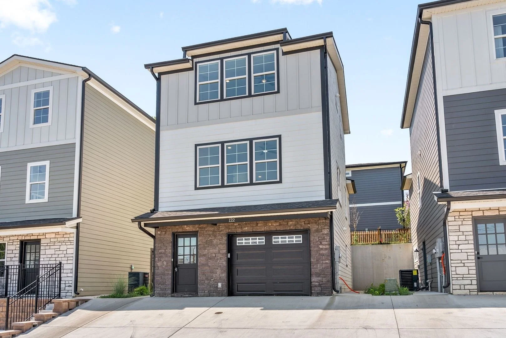 A modern narrow multi-story house with stone and siding exterior, large black garage door, black front door, and multiple windows, in a row of similar style homes under a blue sky.