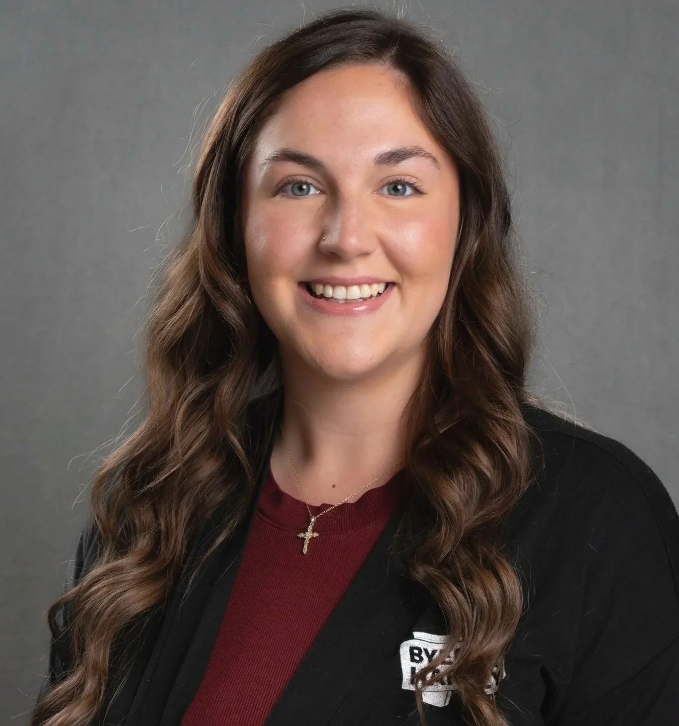 A woman with shoulder-length brown hair, wearing a black blazer and a burgundy top, smiling against a dark gray background.