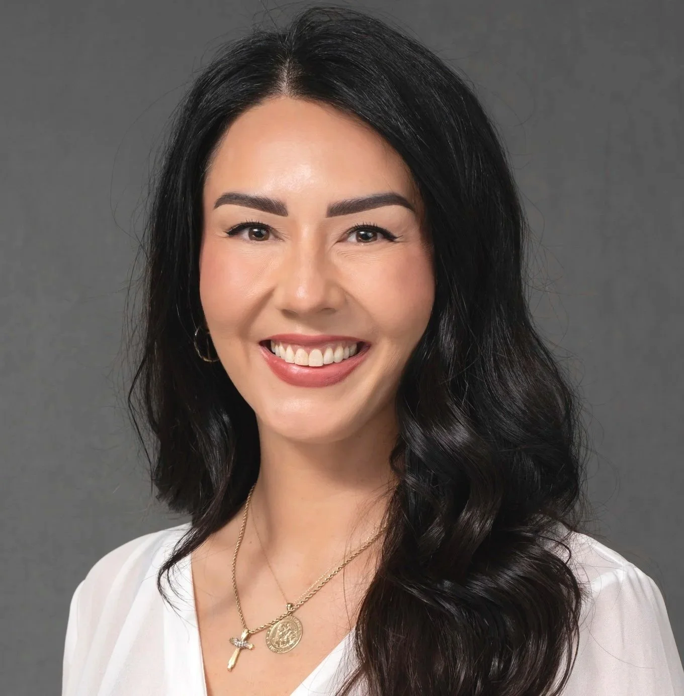 Portrait of a woman with long dark wavy hair, smiling, wearing a black top and black blazer, gold necklace, against a dark background.