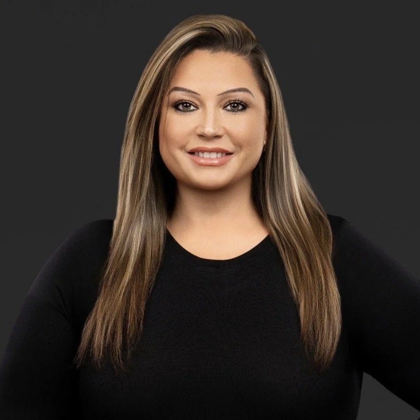 A woman with long brown hair, wearing a black top and a leopard print blazer, smiling against a dark background.