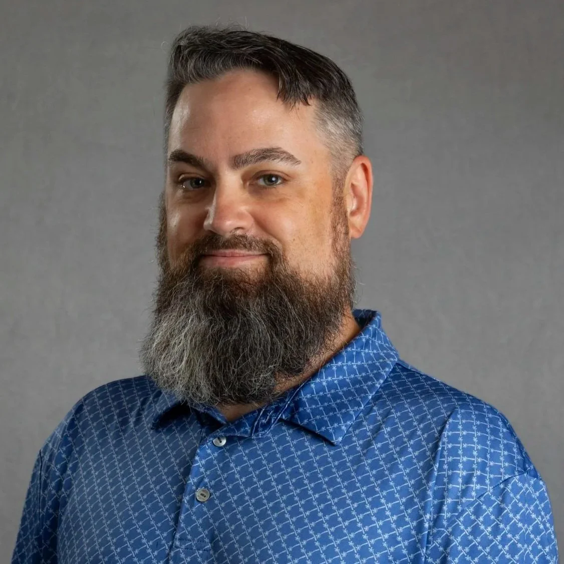 Man with a beard and mustache wearing a blue and white plaid shirt, standing with arms crossed against a dark background.