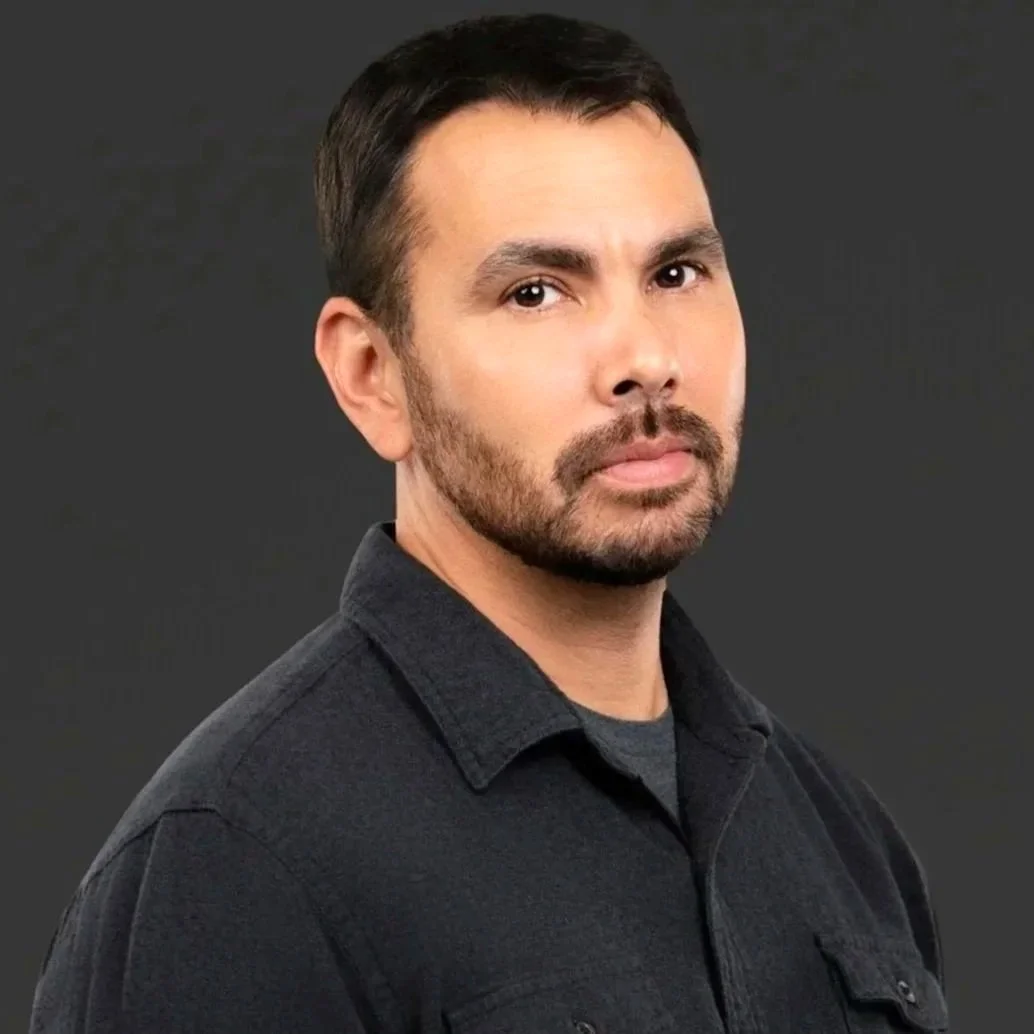 Headshot of a man with dark hair and a beard, wearing a black button-up shirt against a dark background.