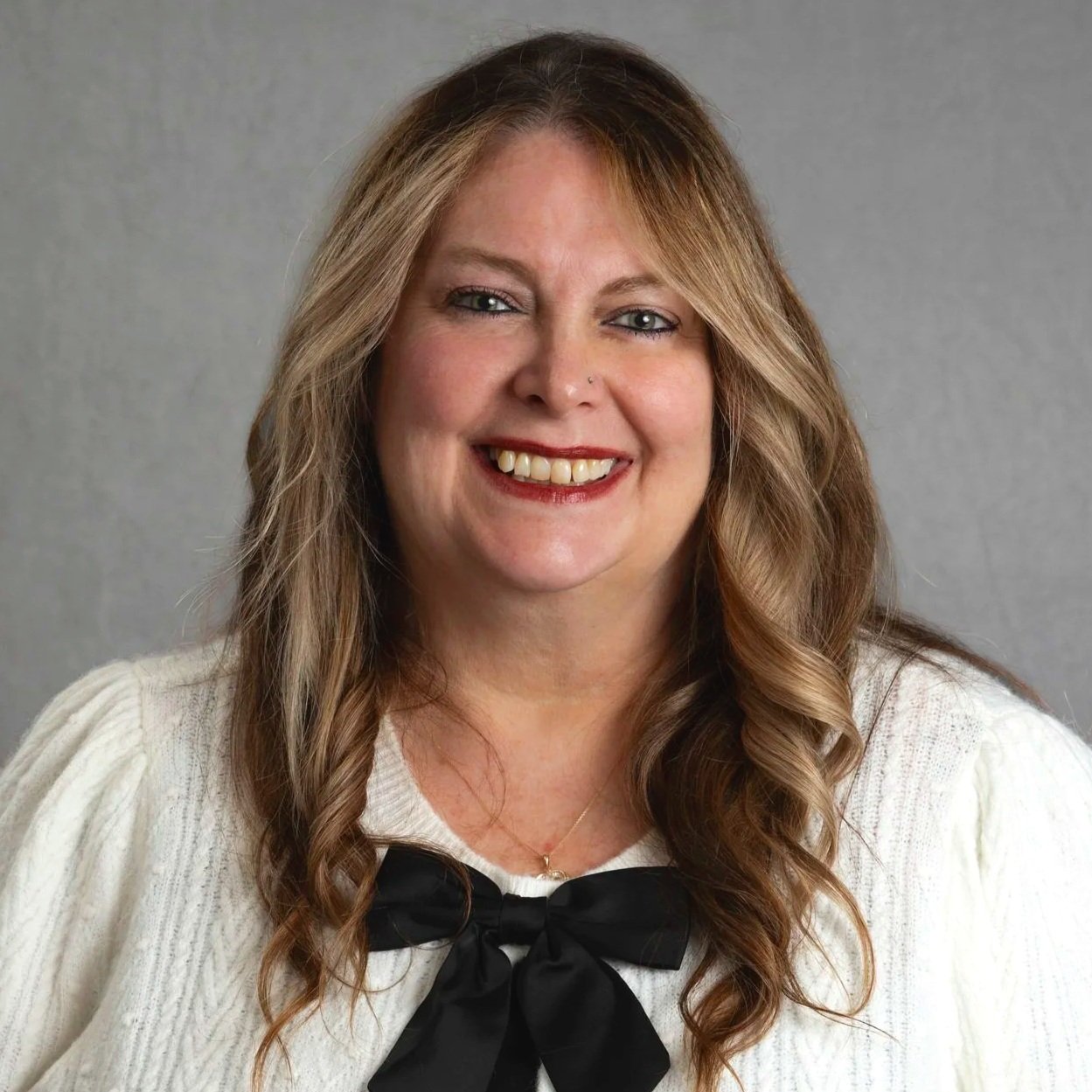 A woman with long wavy blonde hair wearing a black zip-up top and a gold necklace, smiling at the camera against a dark gray background.