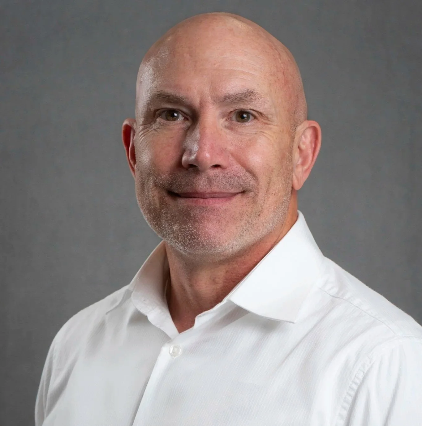 Headshot of a middle-aged man with a bald head and a friendly smile, wearing a white dress shirt against a dark background.