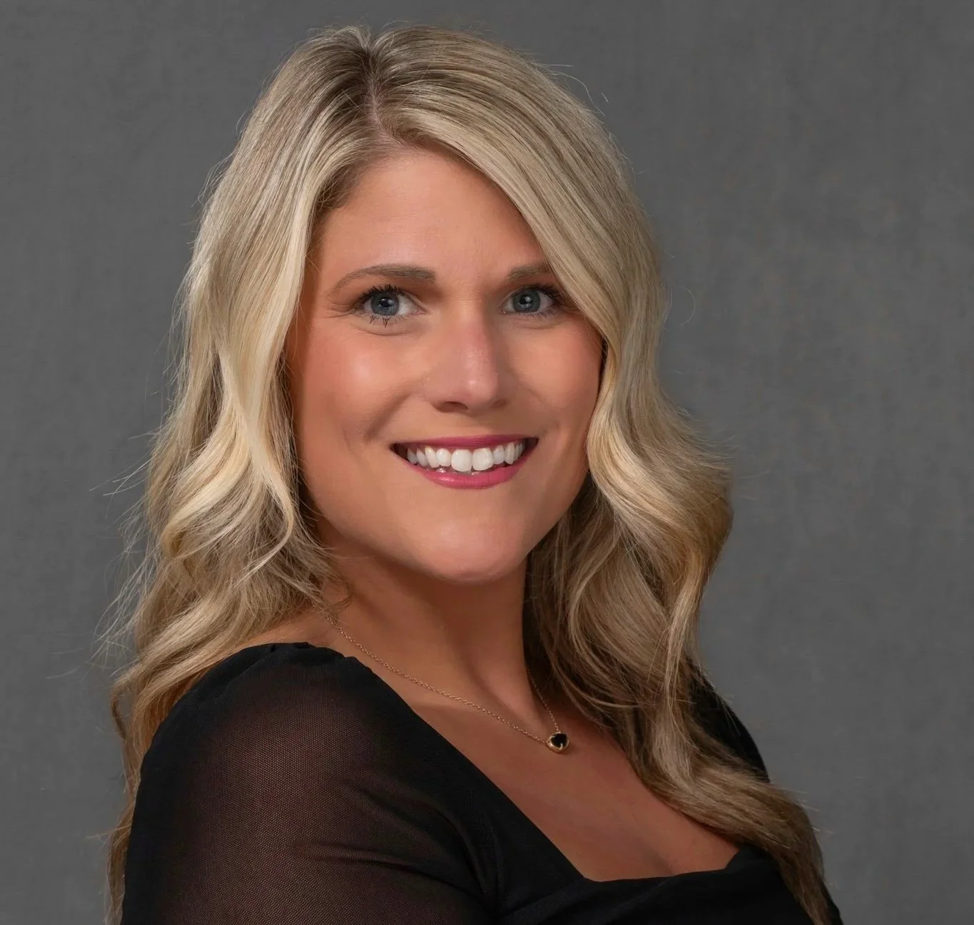 Portrait of a smiling blonde woman with wavy hair, wearing a beige blazer over a black top, against a dark background.