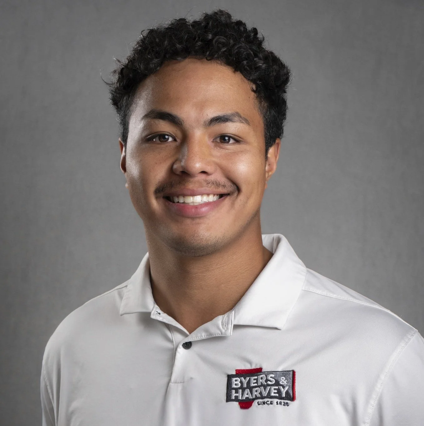 A smiling young man with dark curly hair and light brown skin, wearing an orange striped Nike polo shirt, standing against a dark gray background.