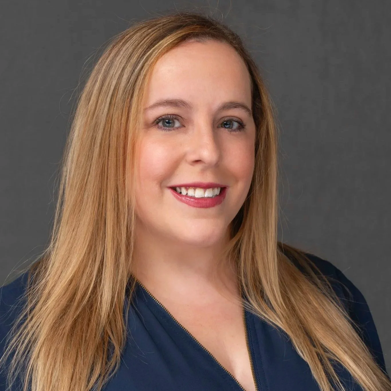 Portrait of a woman with long wavy blonde hair, wearing a dark green shirt, smiling, against a dark gray background.