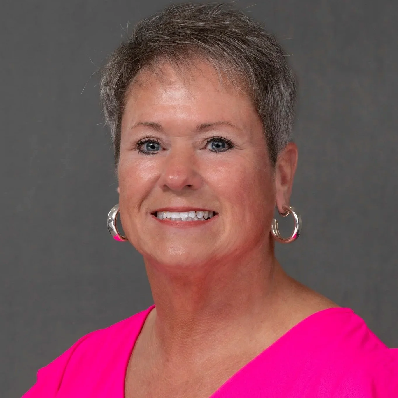 Portrait of a middle-aged woman with short gray hair, wearing a white and gray striped button-up shirt and hoop earrings, smiling against a dark gray background.