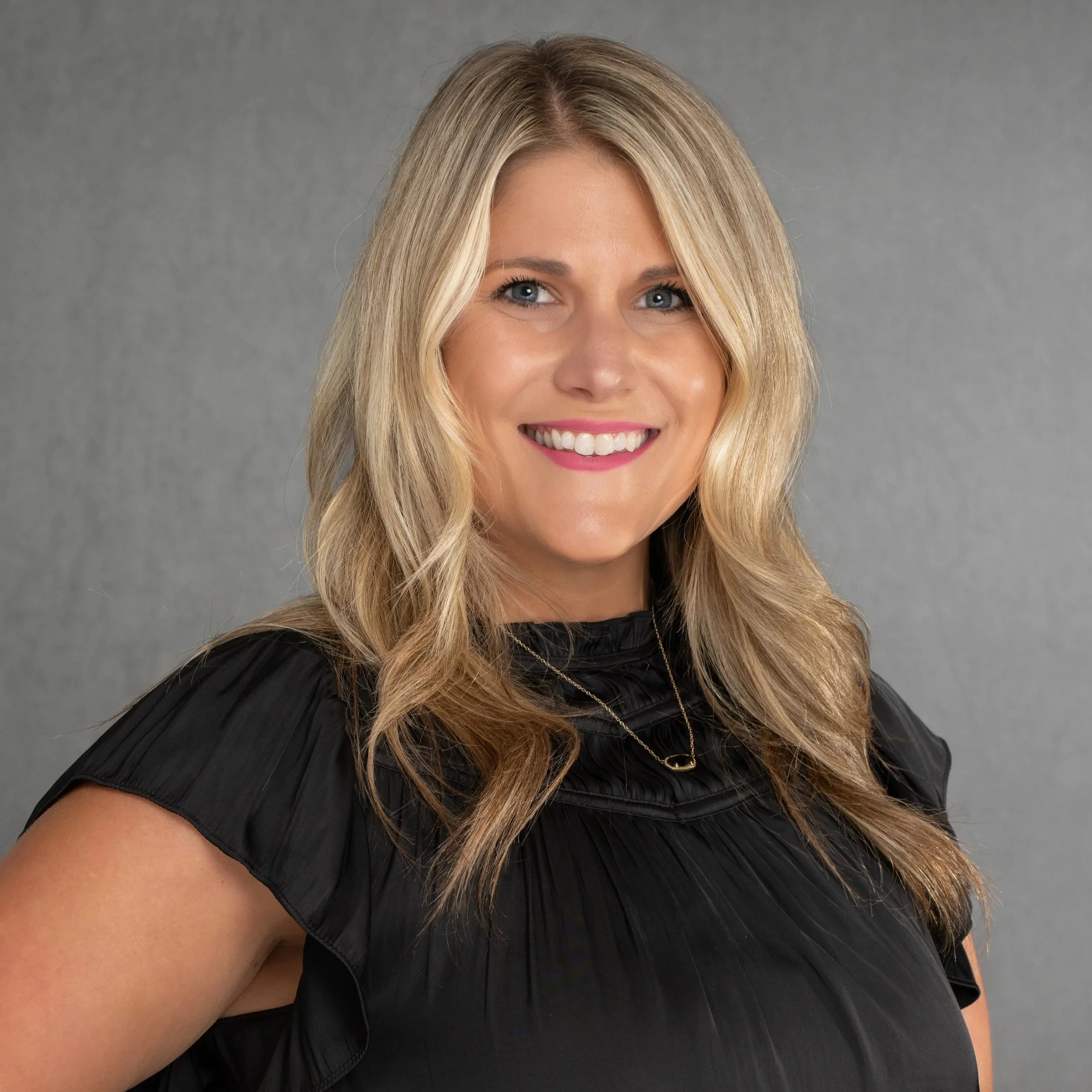A smiling woman with long, wavy blonde hair, wearing a cream-colored blouse with textured dots and a long necklace, posed against a dark gray background.
